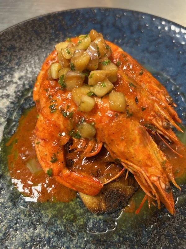 Grilled prawn topped with chopped pickles served on a black plate with sauce.