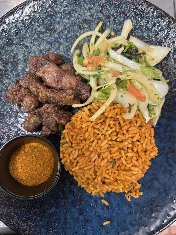 A plate of grilled meat, rice, a vegetable salad, and a small cup of sauce on a blue plate.