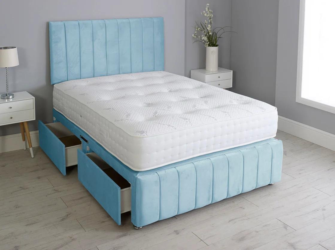 Bedroom with light blue upholstered bed frame and mattress, two white nightstands with drawers, one with a lamp and the other with a tall vase of flowers, light wood flooring, gray walls, and a window.