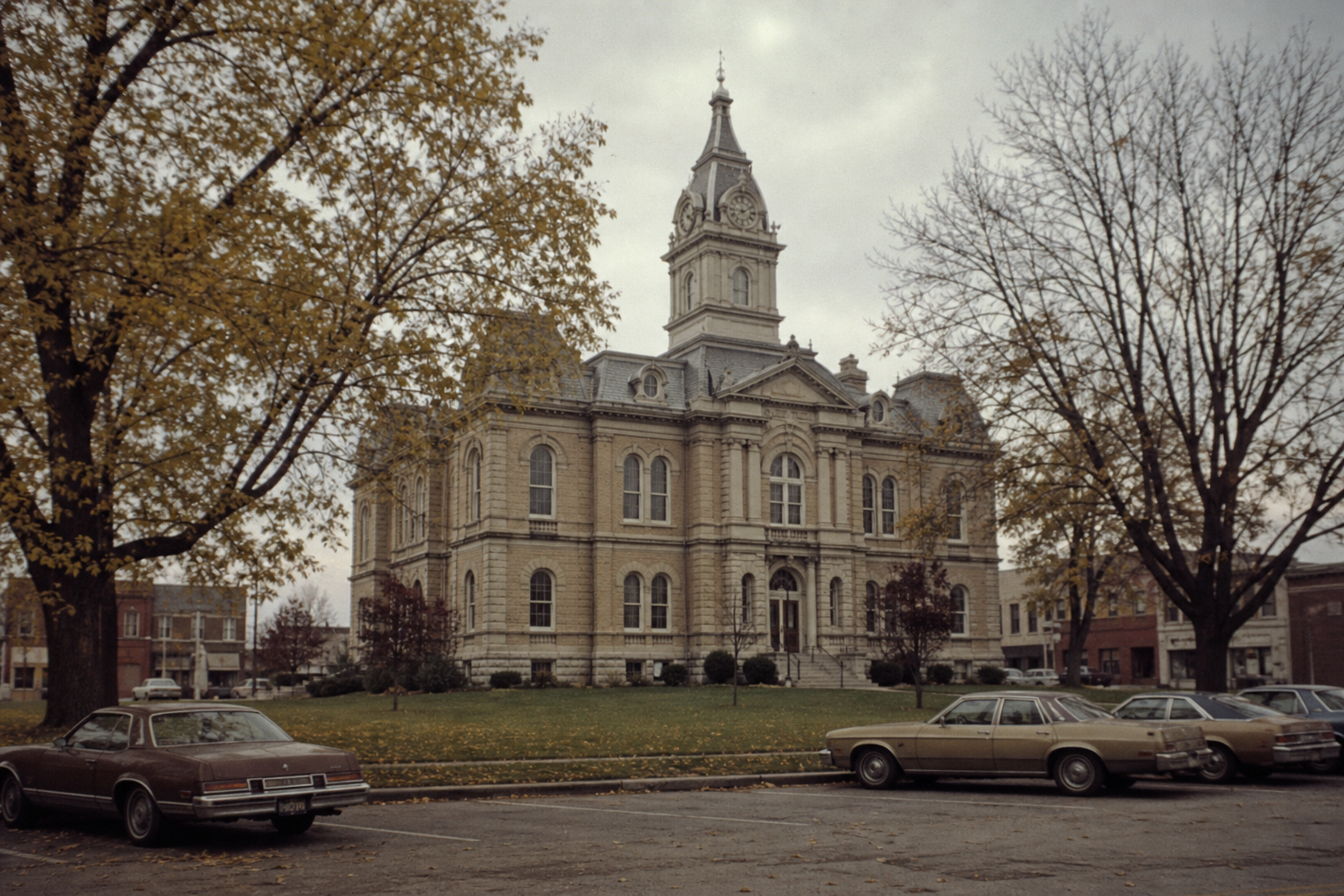Historic courthouse building with a clock tower, surrounded by trees with autumn leaves and parked cars in the foreground.