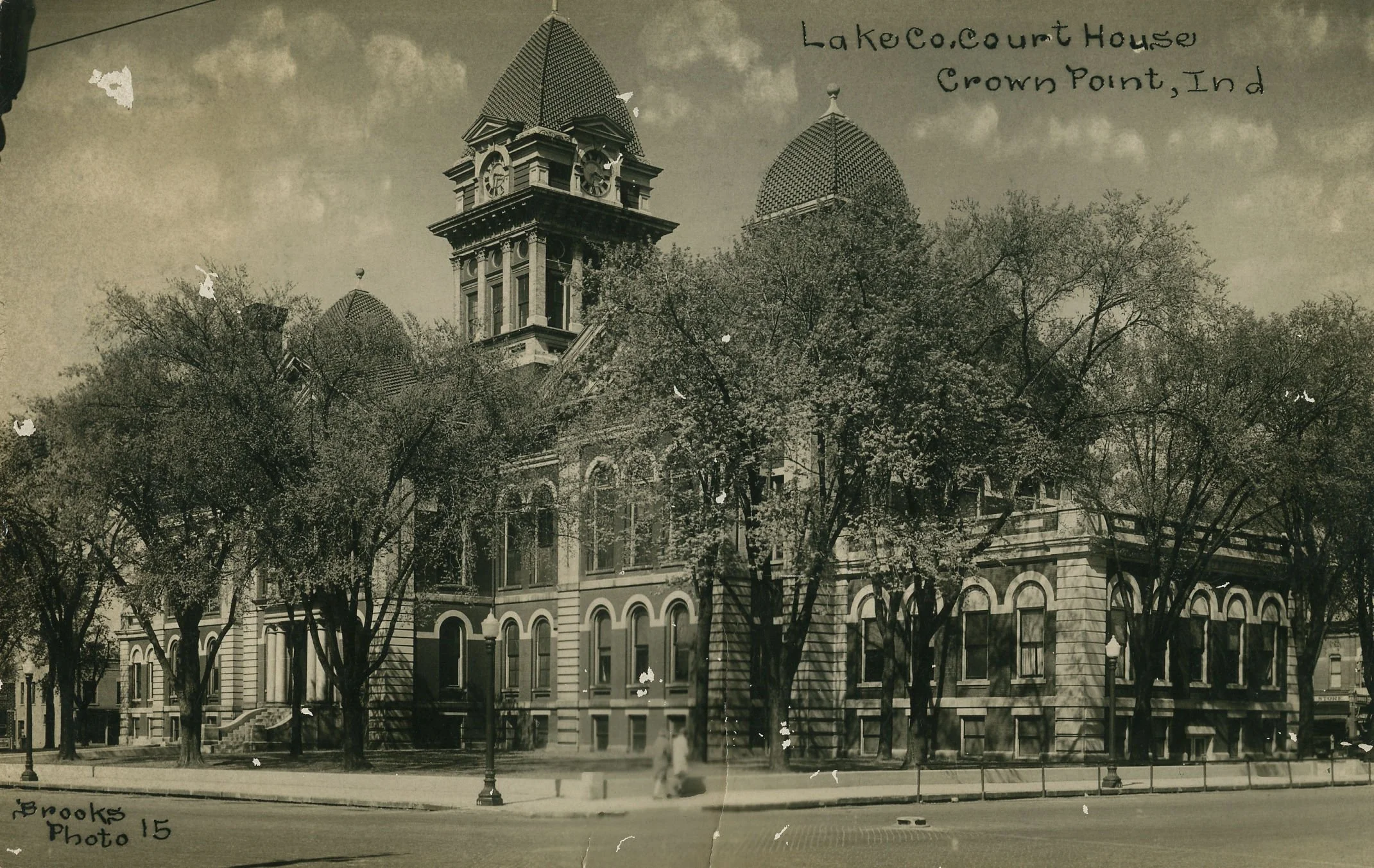 A historic building with two prominent clock towers and a patterned, spiked roof, surrounded by tall trees and street lamps, in Crown Point, Indiana.