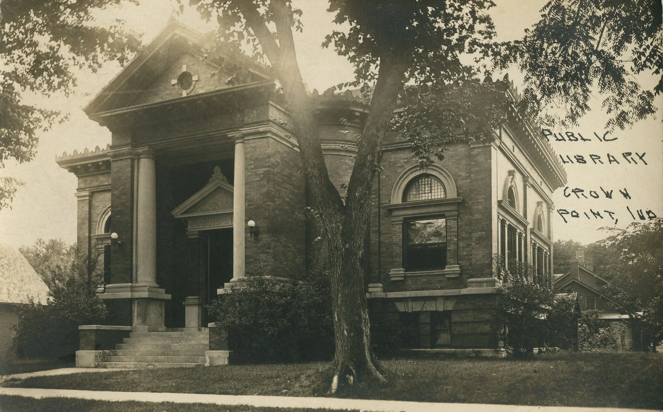 A black and white photograph of a large, historic brick building with Classical architectural features, including columns and decorative elements. There are trees with leaves partially obscuring the building, and a handwritten note on the right side of the image mentioning 'public library, library, crown point, ind'. There are steps leading up to the entrance of the building, and a small part of a garage or house roof is visible in the lower left corner.