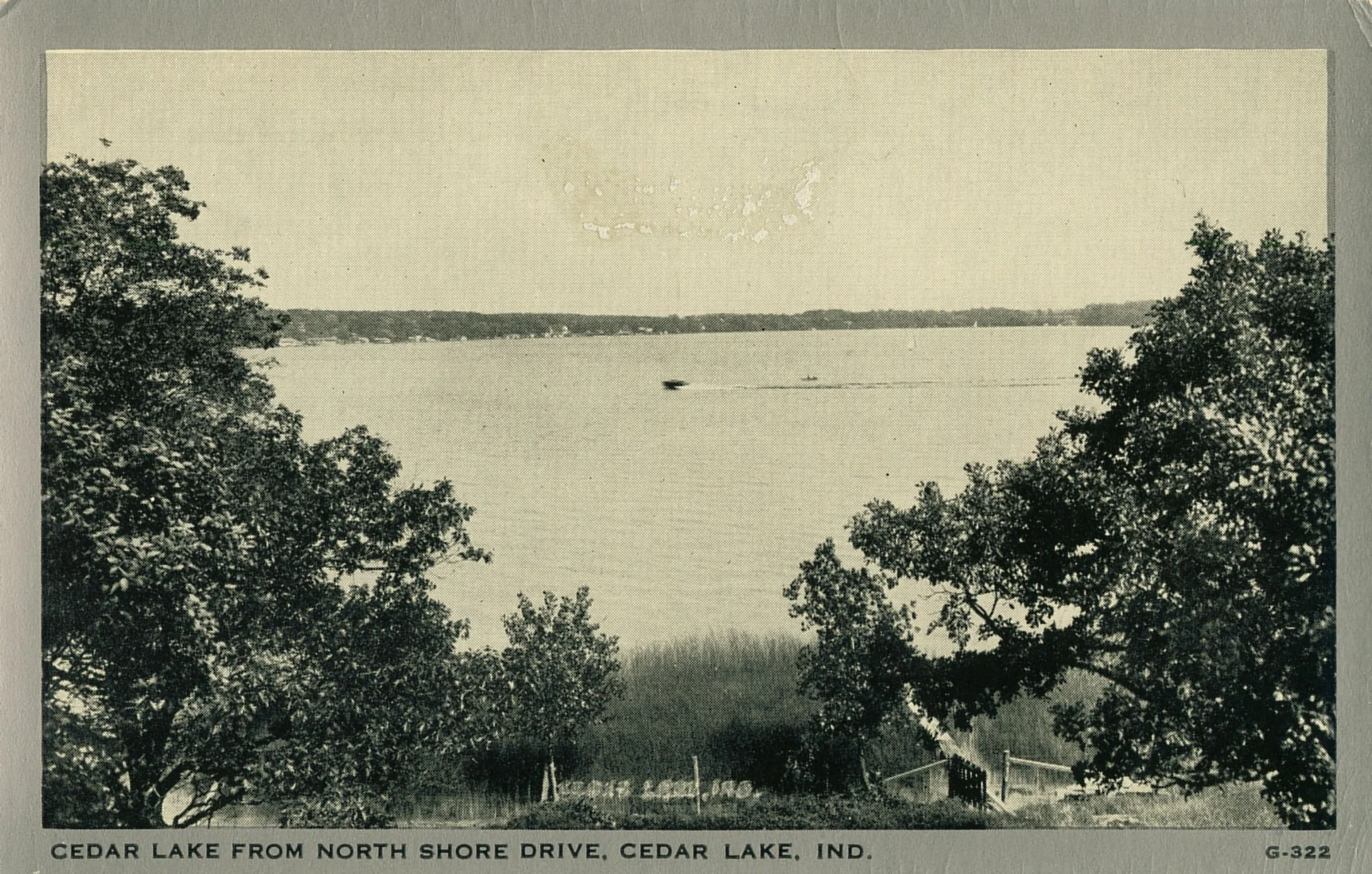 Historic black-and-white view of Cedar Lake from North Shore Drive, with trees in the foreground and a boat on the water.