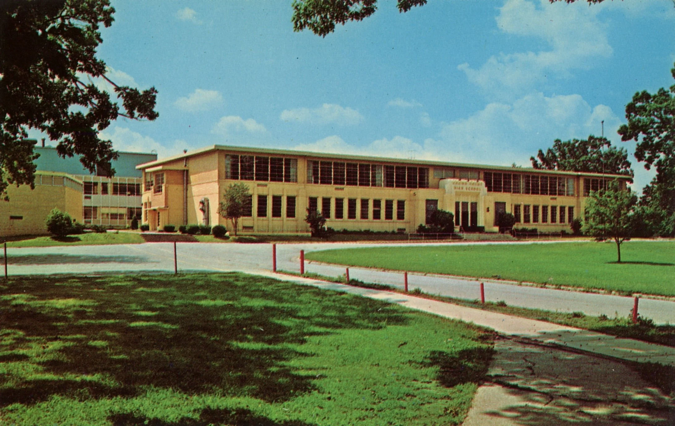 A mid-century school building with large windows, a sign that reads 'High School,' surrounded by green grass, trees, and a clear blue sky.