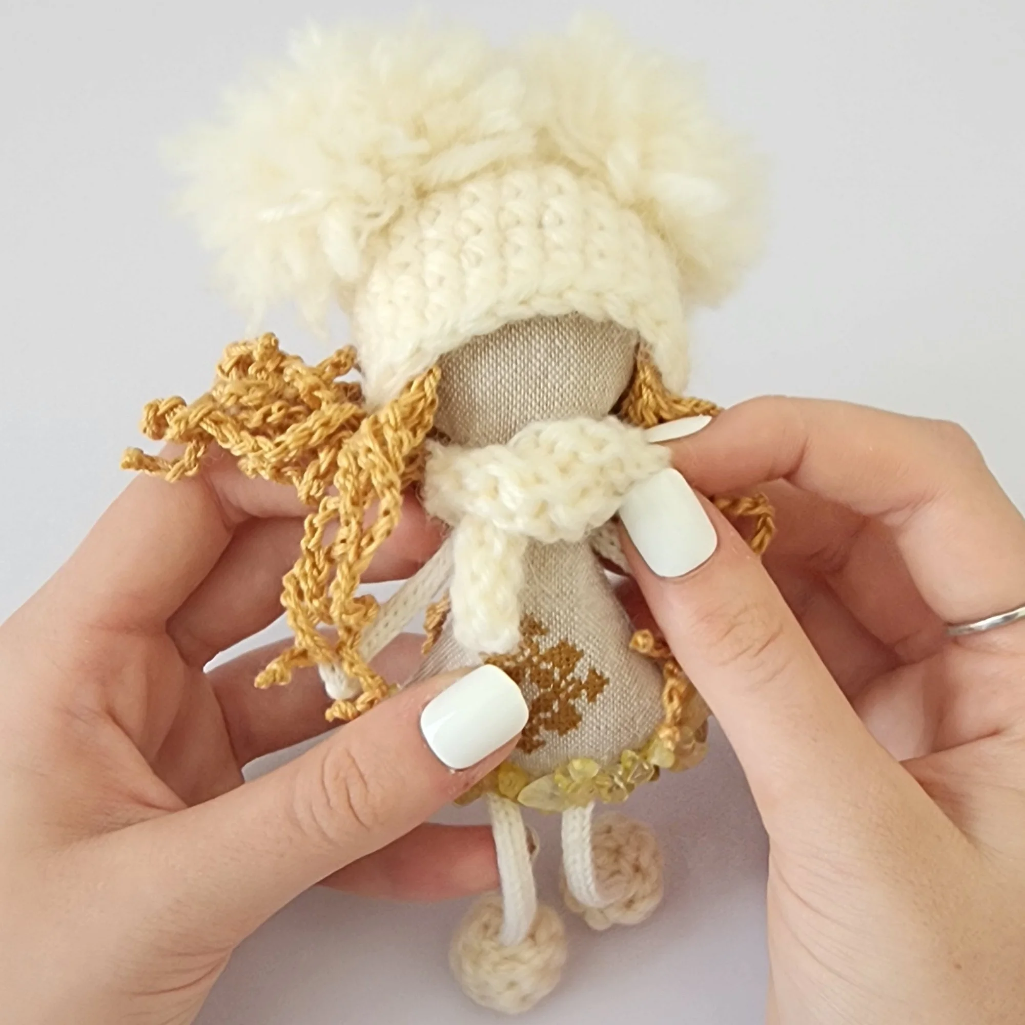Close-up of a handmade cream and gold OMmi held in two hands, showing its textile construction and embroidered ornament.