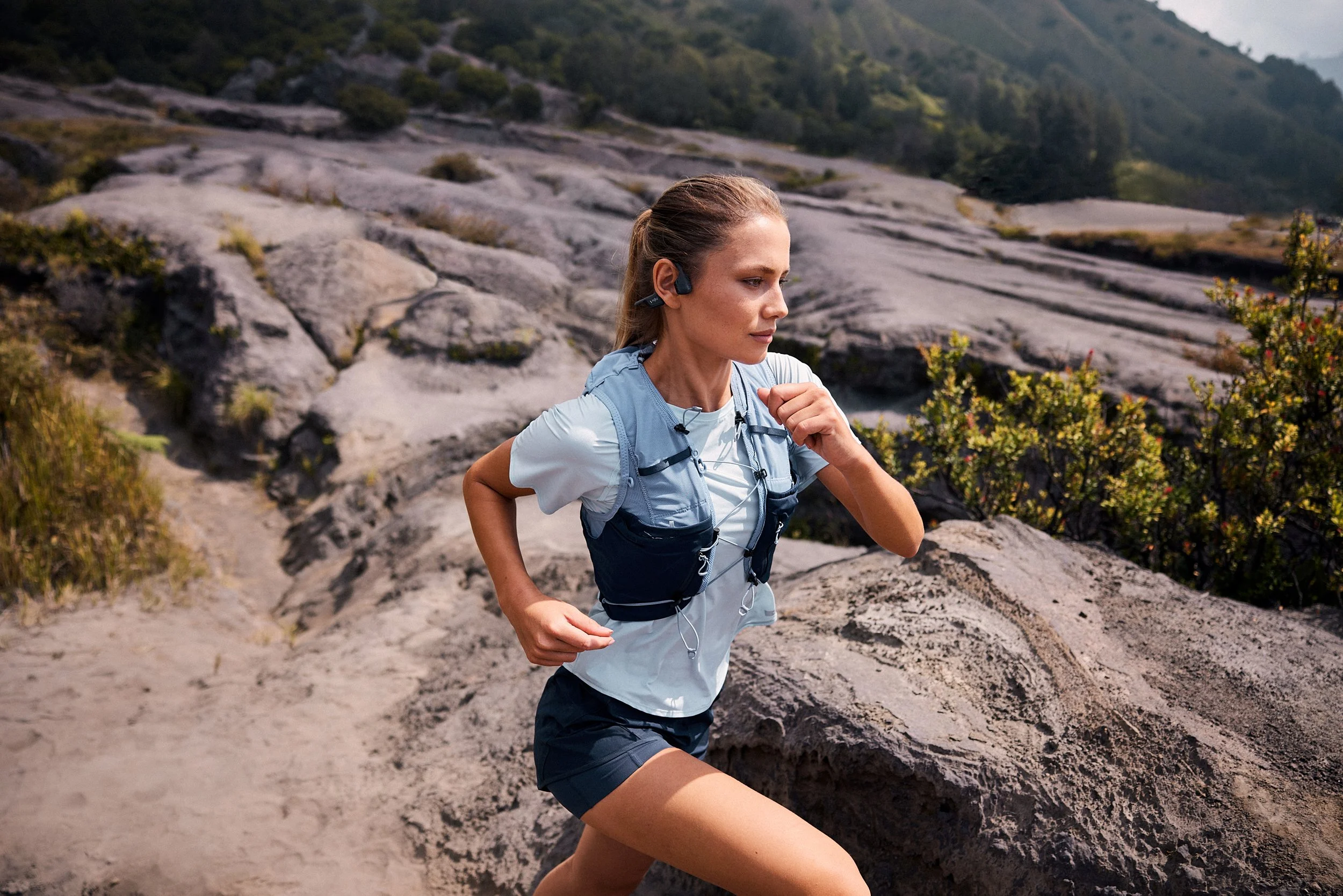 A woman running along a rocky trail in a mountainous area, wearing a light blue shirt, black shorts, and a blue hydration pack, with a mountain landscape in the background.