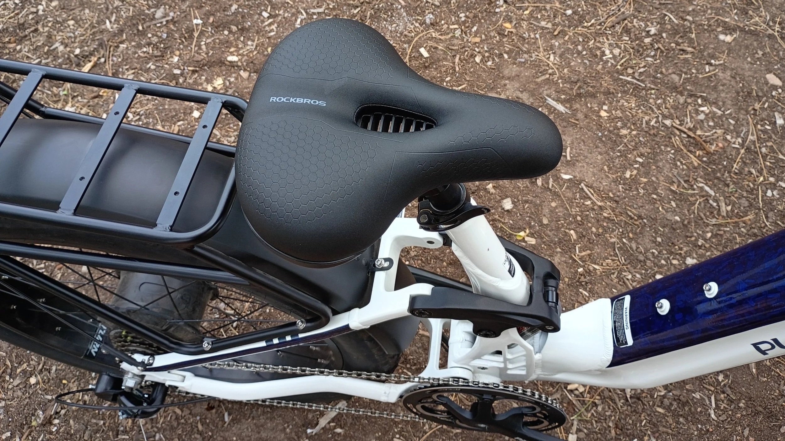 View of a bicycle saddle marked 'ROCKBROS' on a white bike frame with front suspension, located on dirt ground with small rocks and twigs.