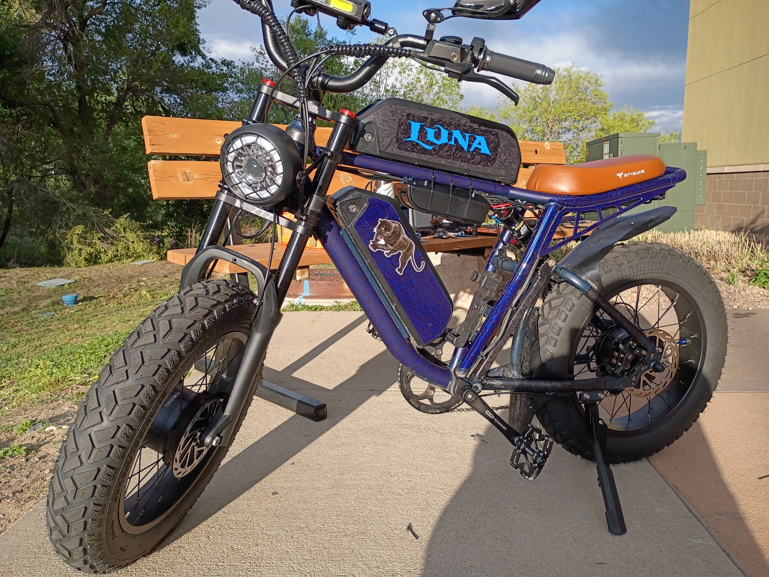 Close-up of a blue electric mountain bike with large tires, a headlamp, and a black and blue frame, parked on a concrete surface with a wooden bench and trees in the background.