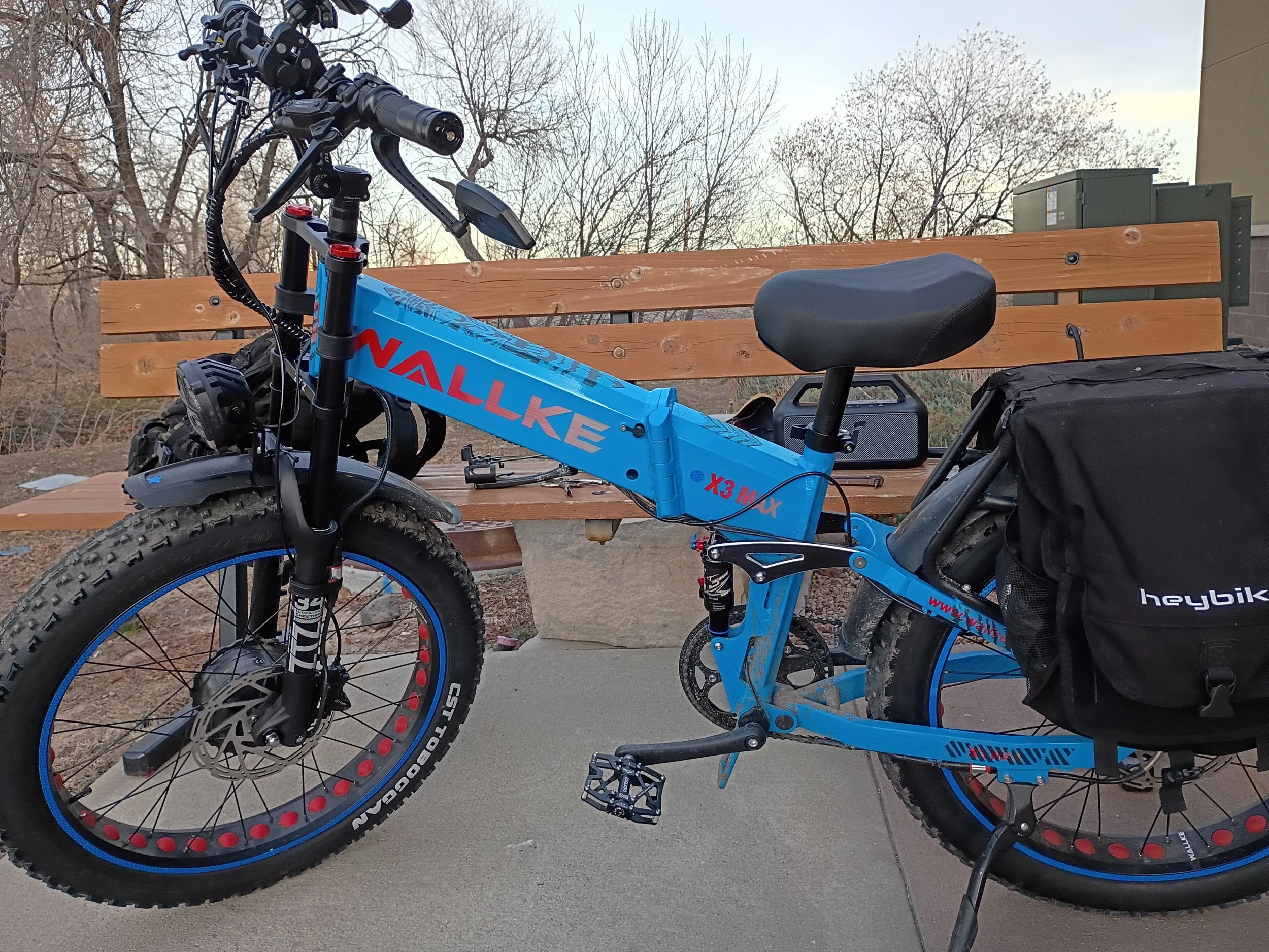 A blue mountain bike with black accents and large knobby tires parked next to a wooden park bench. In the background, leafless trees and utility boxes are visible against a light sky.