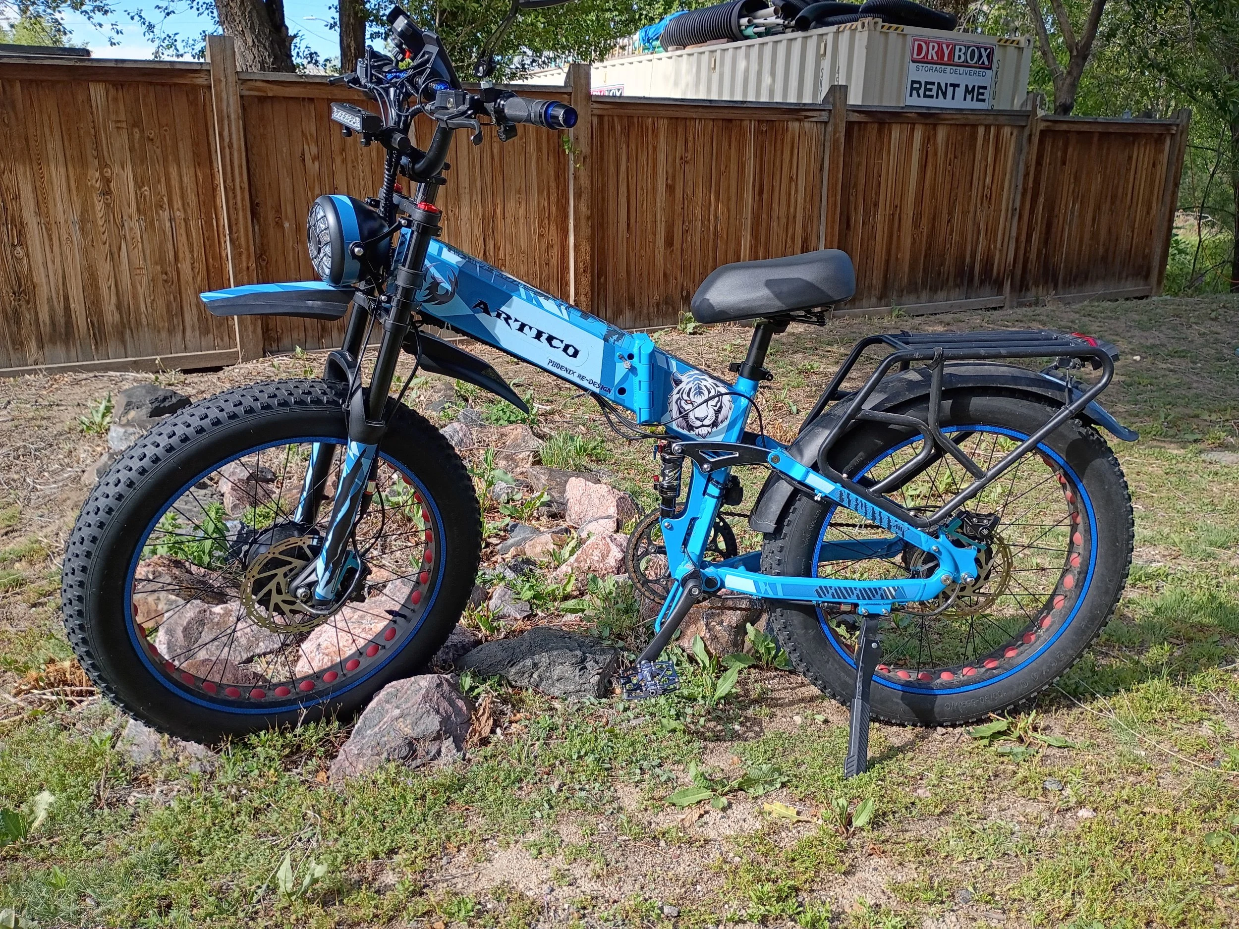 Blue electric mountain bike with fat tires, parked on rocky ground in front of a wooden fence, with a storage container and trees in the background.