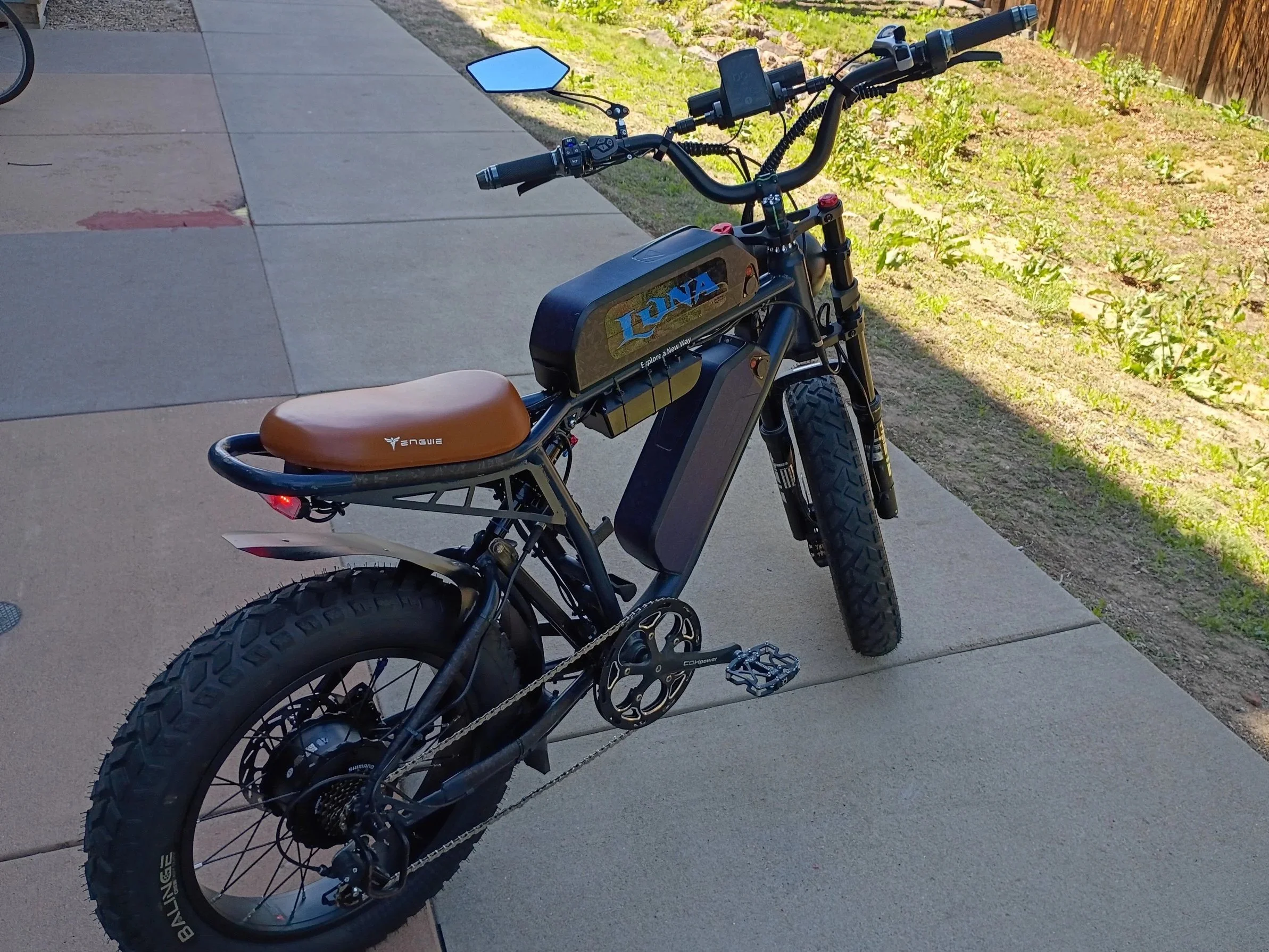 An e-bike with a brown leather seat and black frame, parked on a sidewalk near grass and a wooden fence, with a large black battery labeled 'LUNA' and a small blue side mirror on the handlebar.