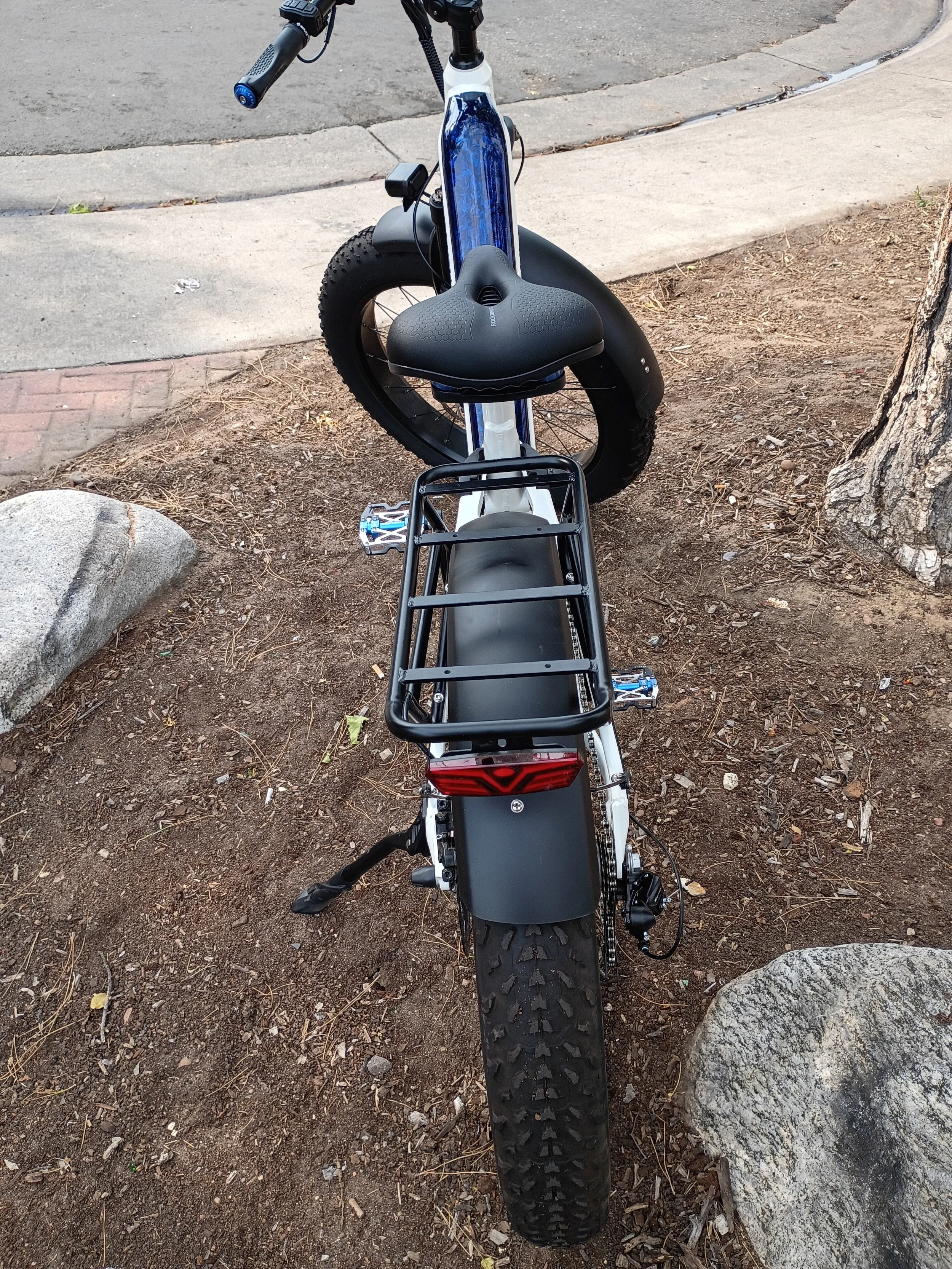 A black electric bicycle parked on a dirt patch near a sidewalk, with a rear rack and knobby tires, viewed from the front.