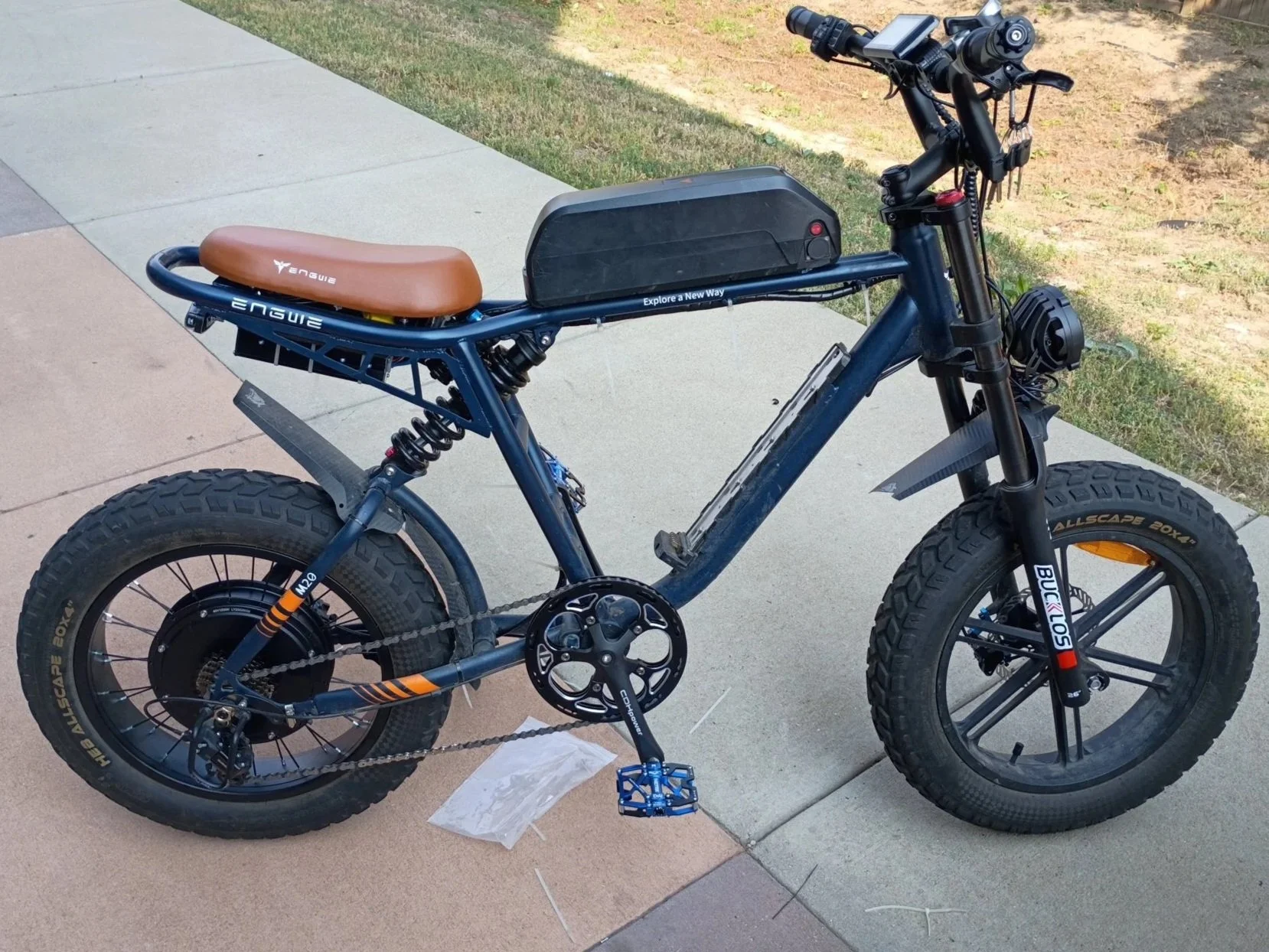 An electric mountain bike with large off-road tires parked on a sidewalk next to grass. The bike has a brown saddle, a black frame with brand labels, a battery on top of the frame, and front suspension forks.