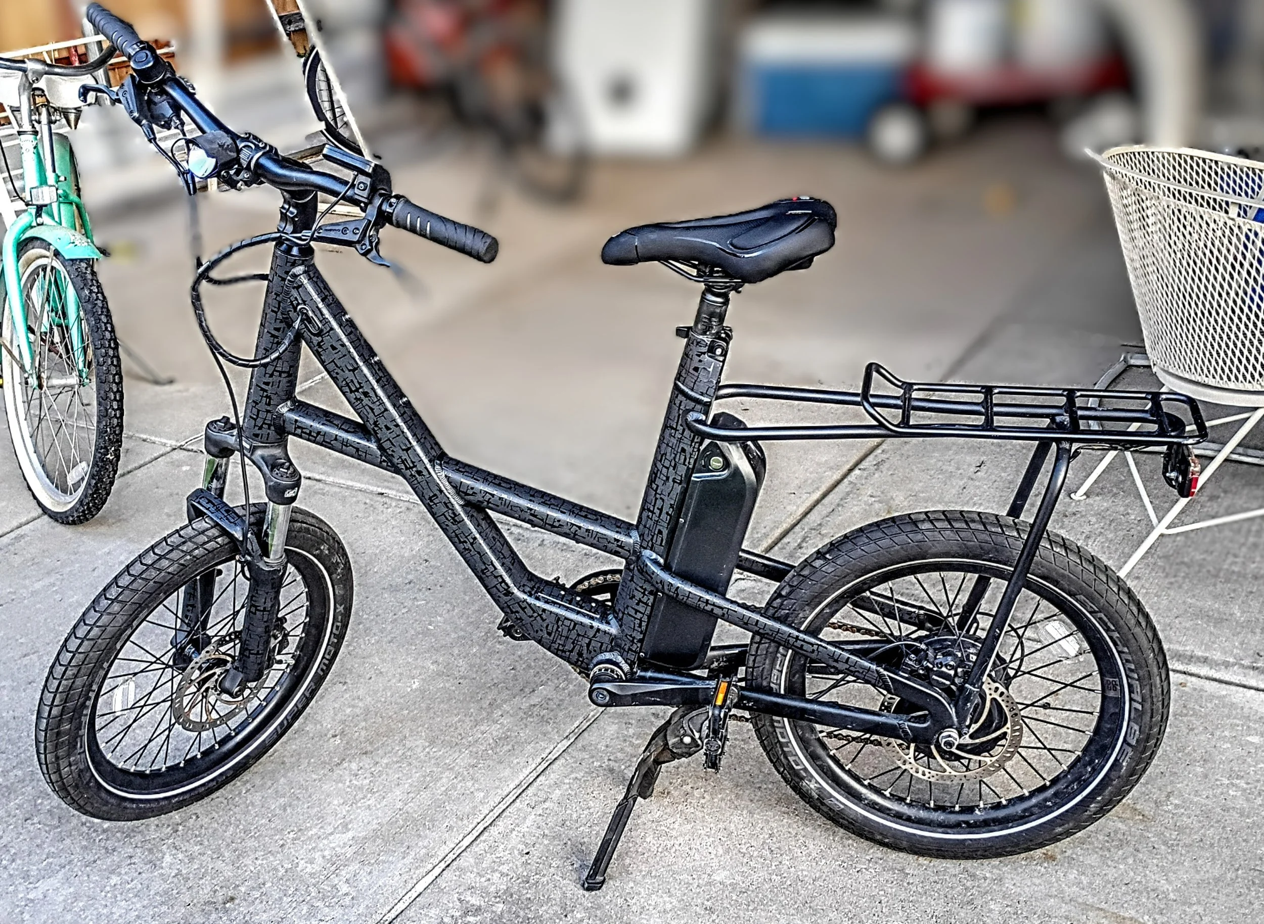 Two bicycles, one black and one mint green, parked on a concrete sidewalk. The black bicycle has a basket attachment, and the mint green bicycle is partially visible in the background.