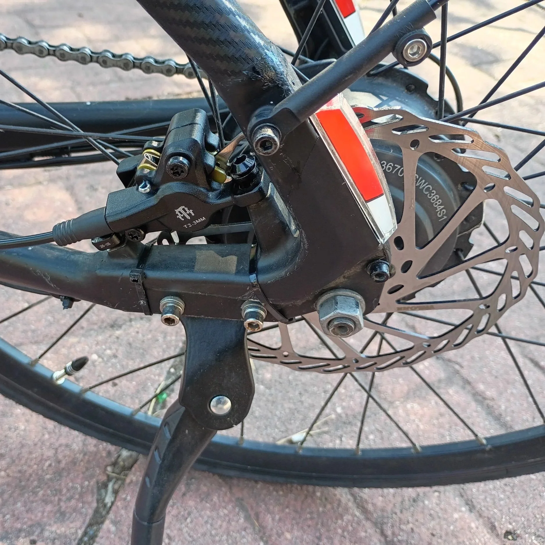 Close-up of the front wheel of an electric bicycle, showing the disc brake, spokes, and part of the frame, with a background of pavement.