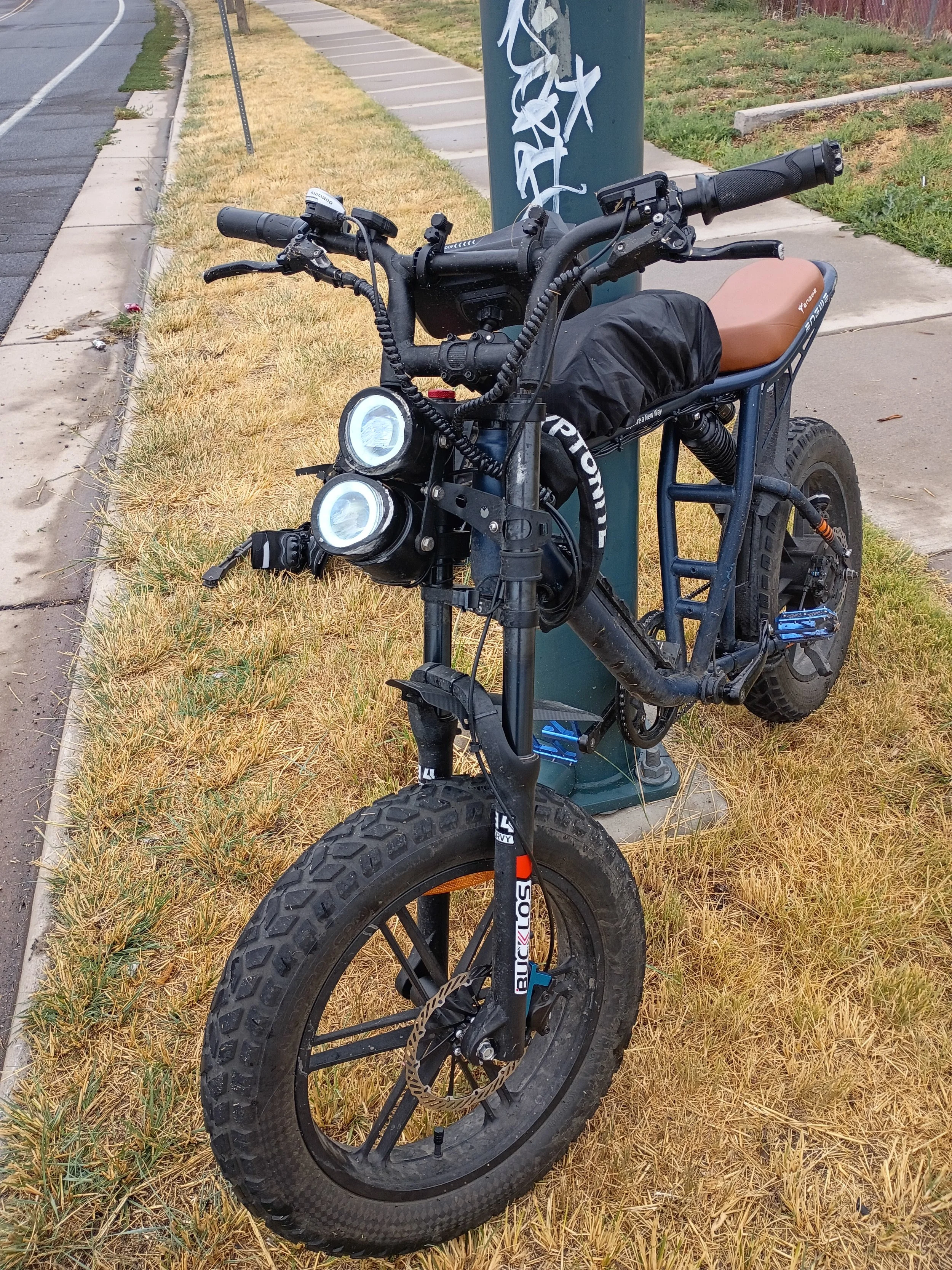 An electric bike parked on the grass beside the sidewalk, locked to a green utility pole with white graffiti. The bike has a tan seat, black frame, and large tires.