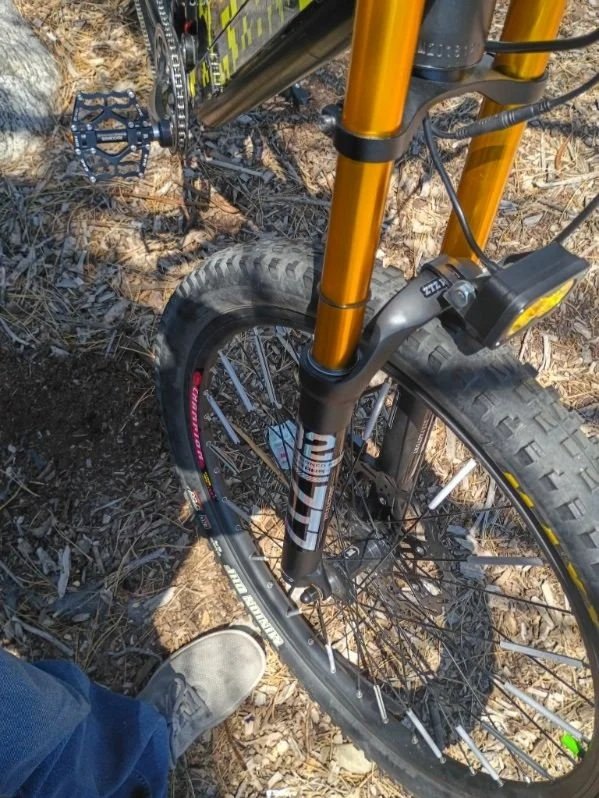 Close-up of a mountain bike's front wheel, fork, and suspension on a dirt trail, with a person's foot in gray shoe visible in the lower left corner.