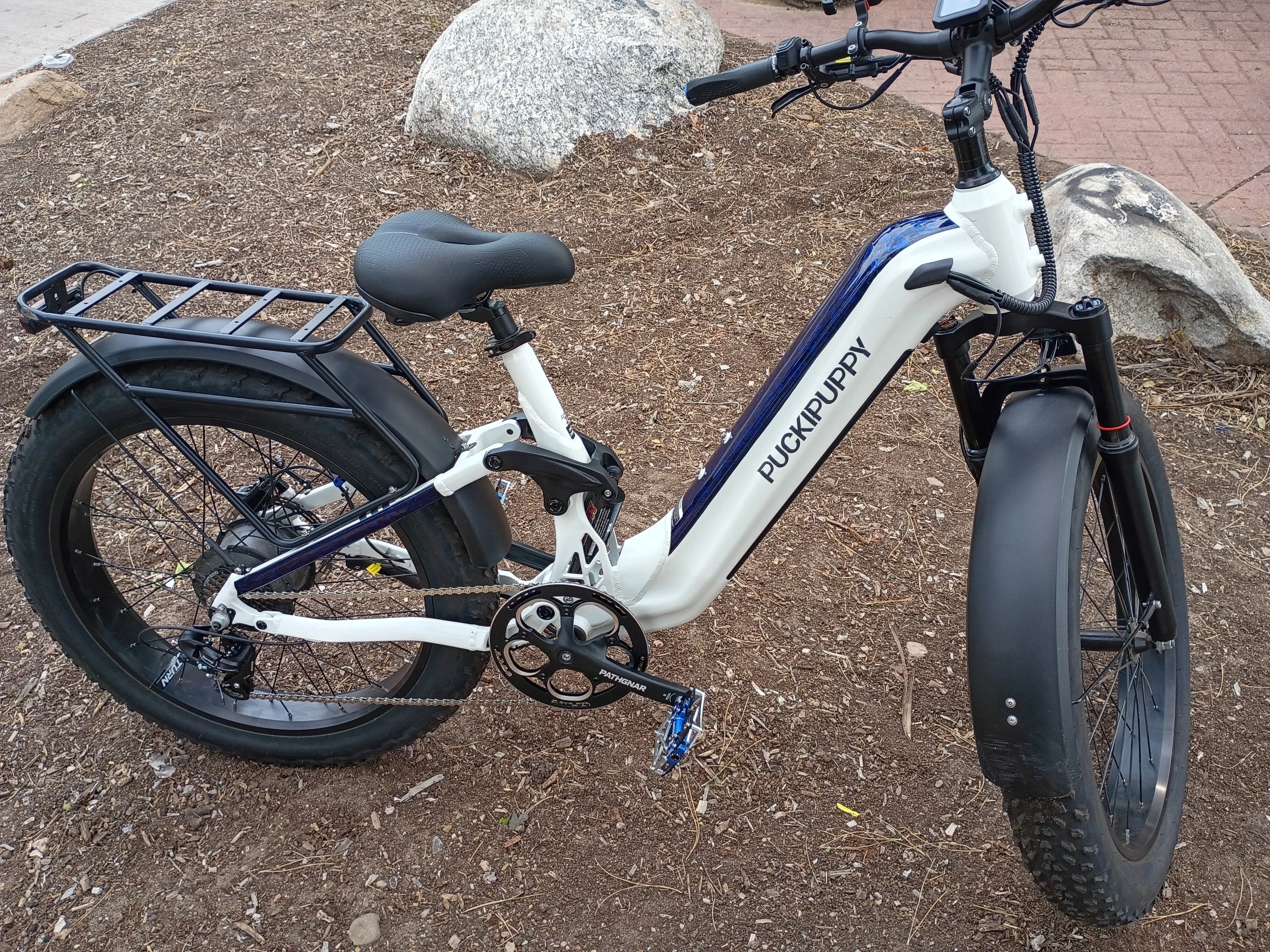 A white and black electric bicycle on a dirt patch next to a brick sidewalk, with rocks in the background.