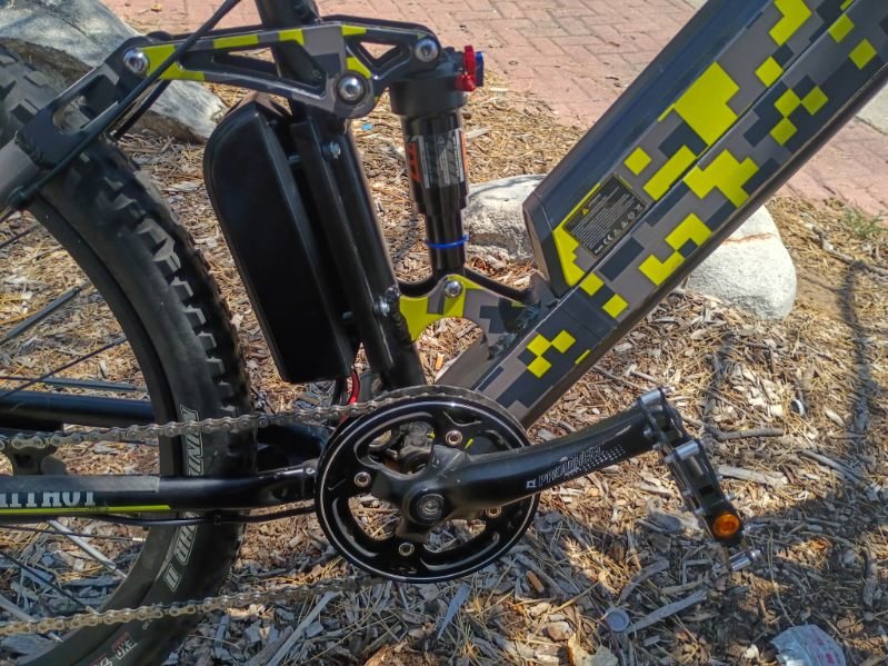 Close-up of a mountain bike's frame showing the crankset, chain, pedal, and rear suspension, with a rocky outdoor ground background.