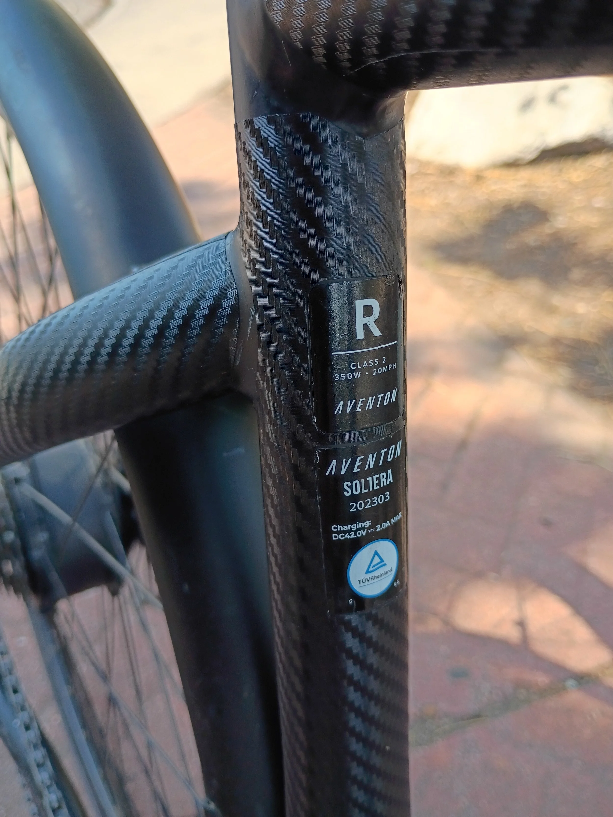 Close-up of a bicycle front fork with a black carbon fiber finish, showing a label indicating the model as Aventon Soltera, with details about the motor and battery charging.