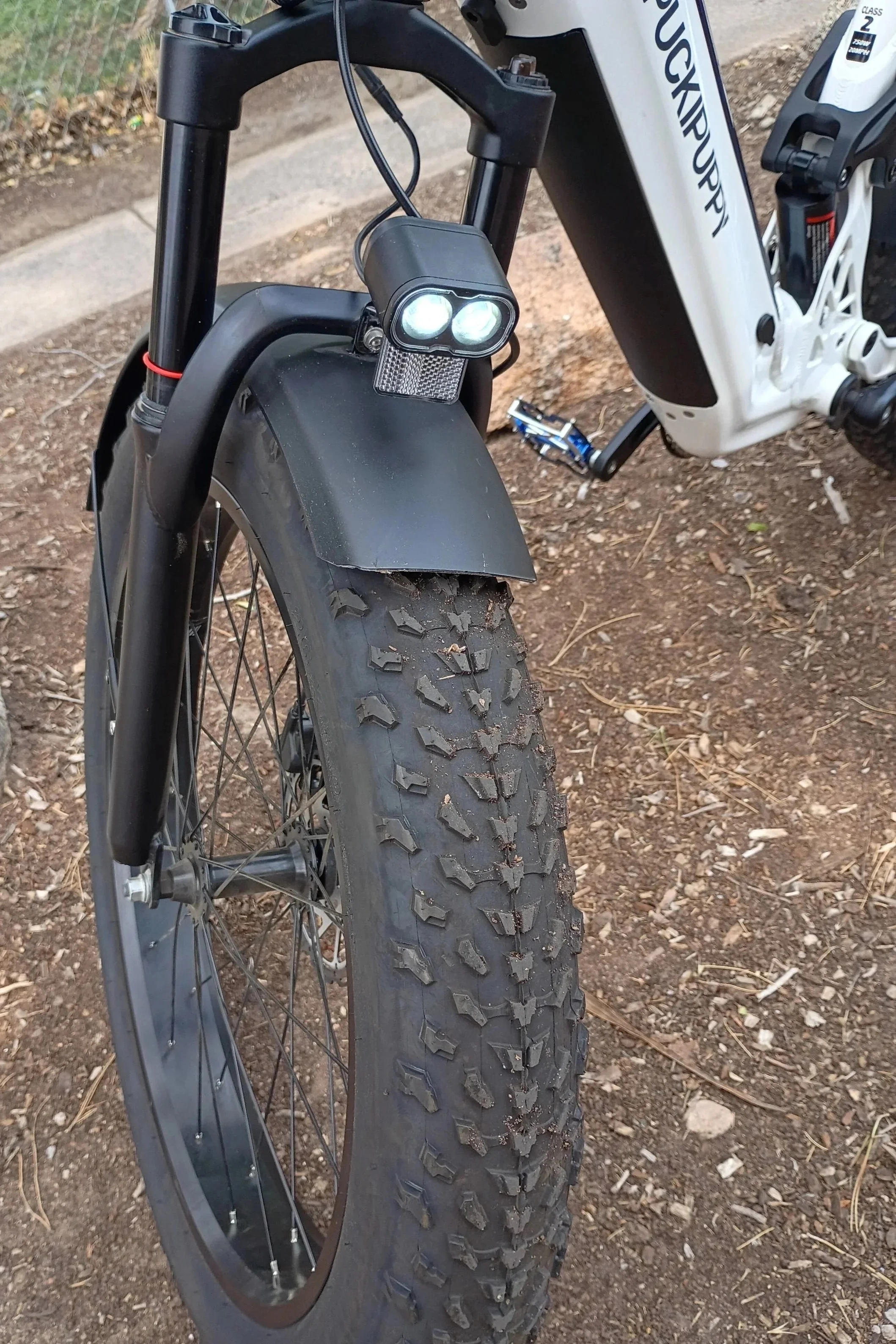 Close-up of the front part of an electric mountain bike, showing the front wheel, tire with mud, suspension fork, and front light, on dirt ground.