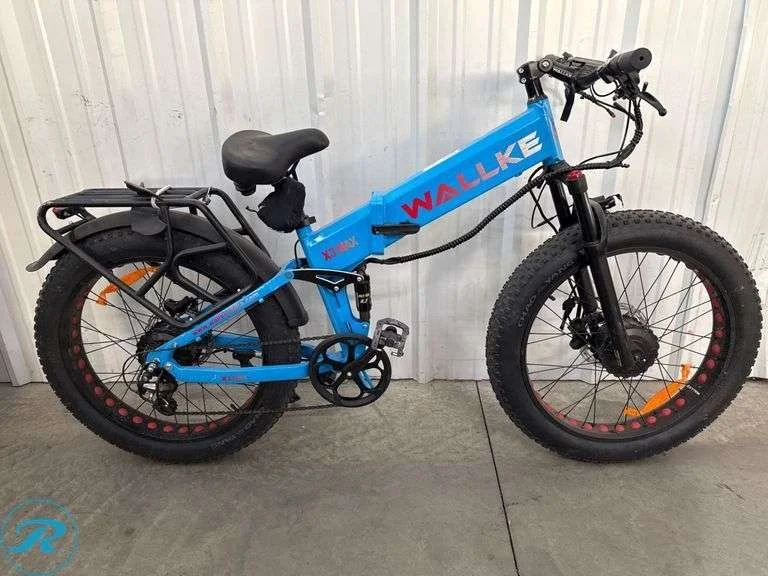 Blue fat tire electric bike with black seat, black rear rack, and front disc brake, parked against a white wall.