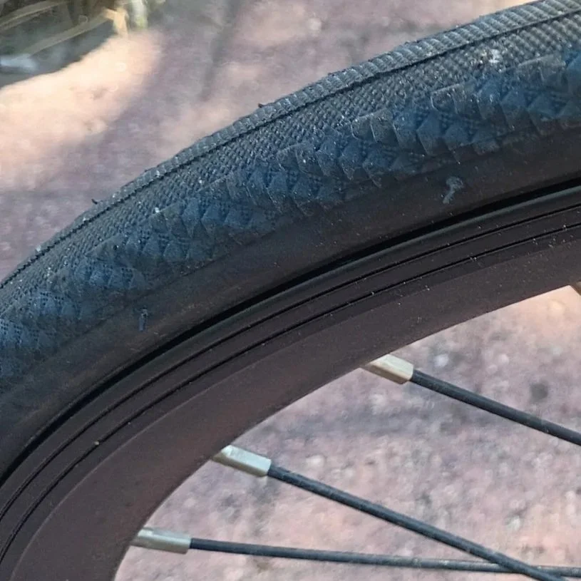 Close-up of a bicycle tire with a worn tread and spokes visible, with a gravel surface in the background.