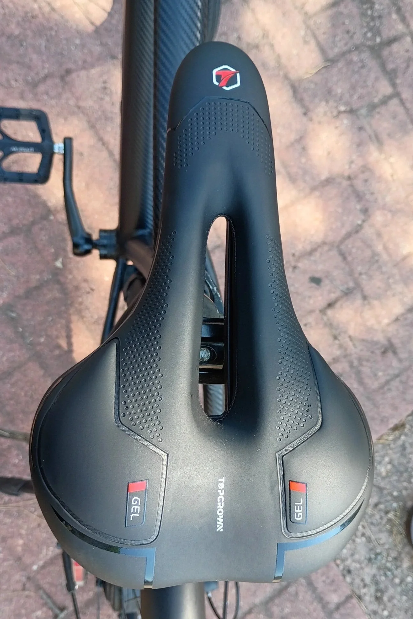 Top-down view of a black bicycle saddle with a logo and gel padding, mounted on a bike on a brick-paved surface.