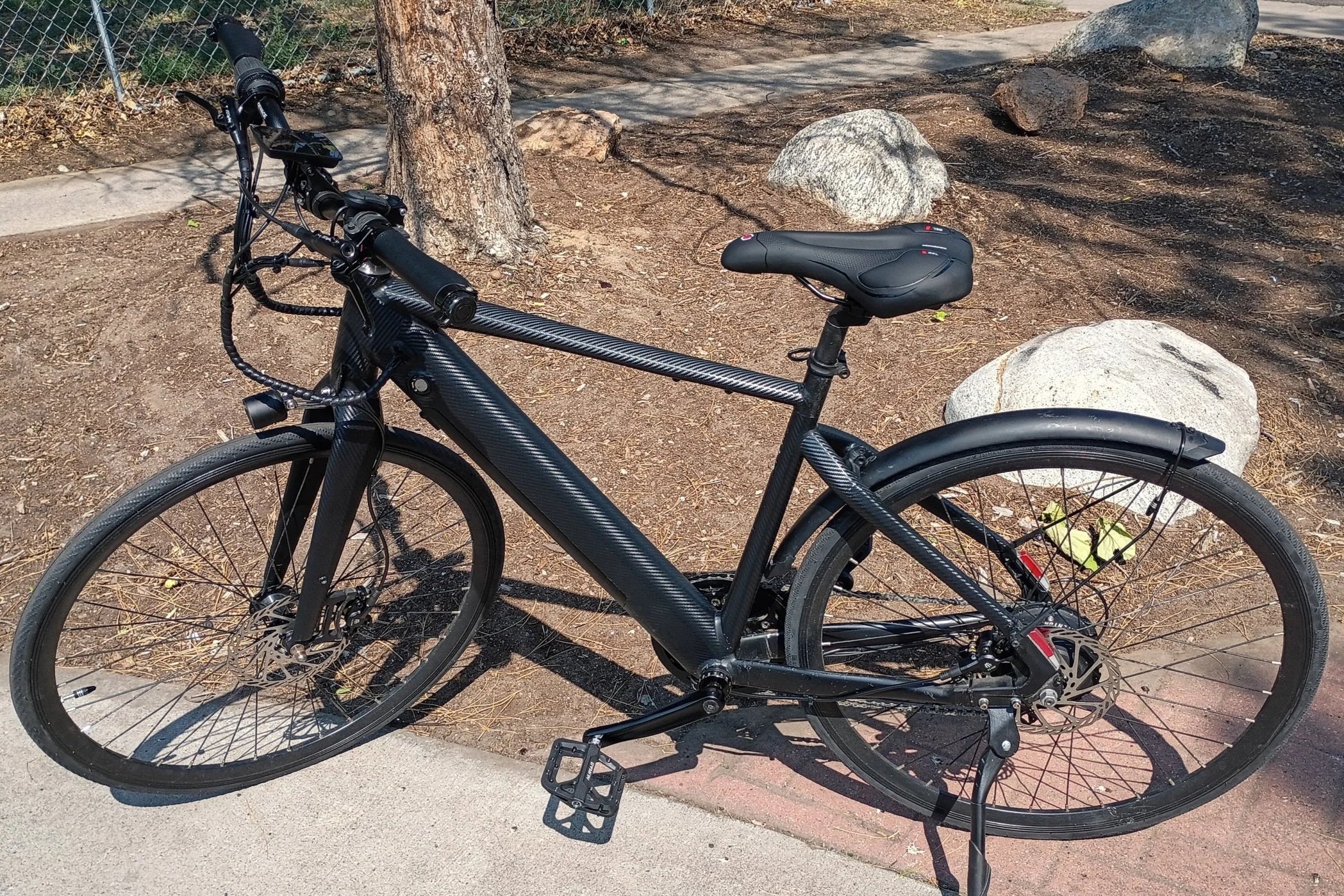 A black carbon fiber mountain bike with a padded saddle, flat handlebars, and disc brakes parked on a sidewalk next to a tree and large rocks, with a chain-link fence in the background.