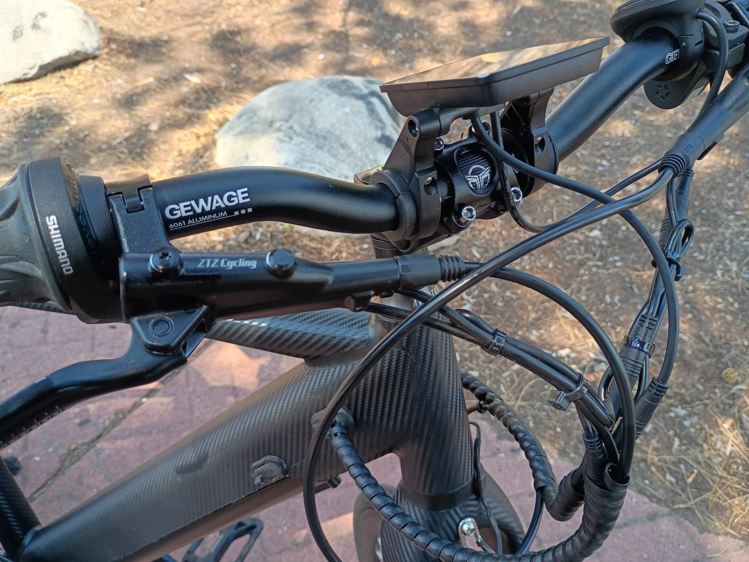 Close-up view of a black electric bicycle handlebar with a mounted screen, cables, and branding that reads GEWAGE, ZTZ Cycling, and Shimano. The background shows dirt and scattered rocks.
