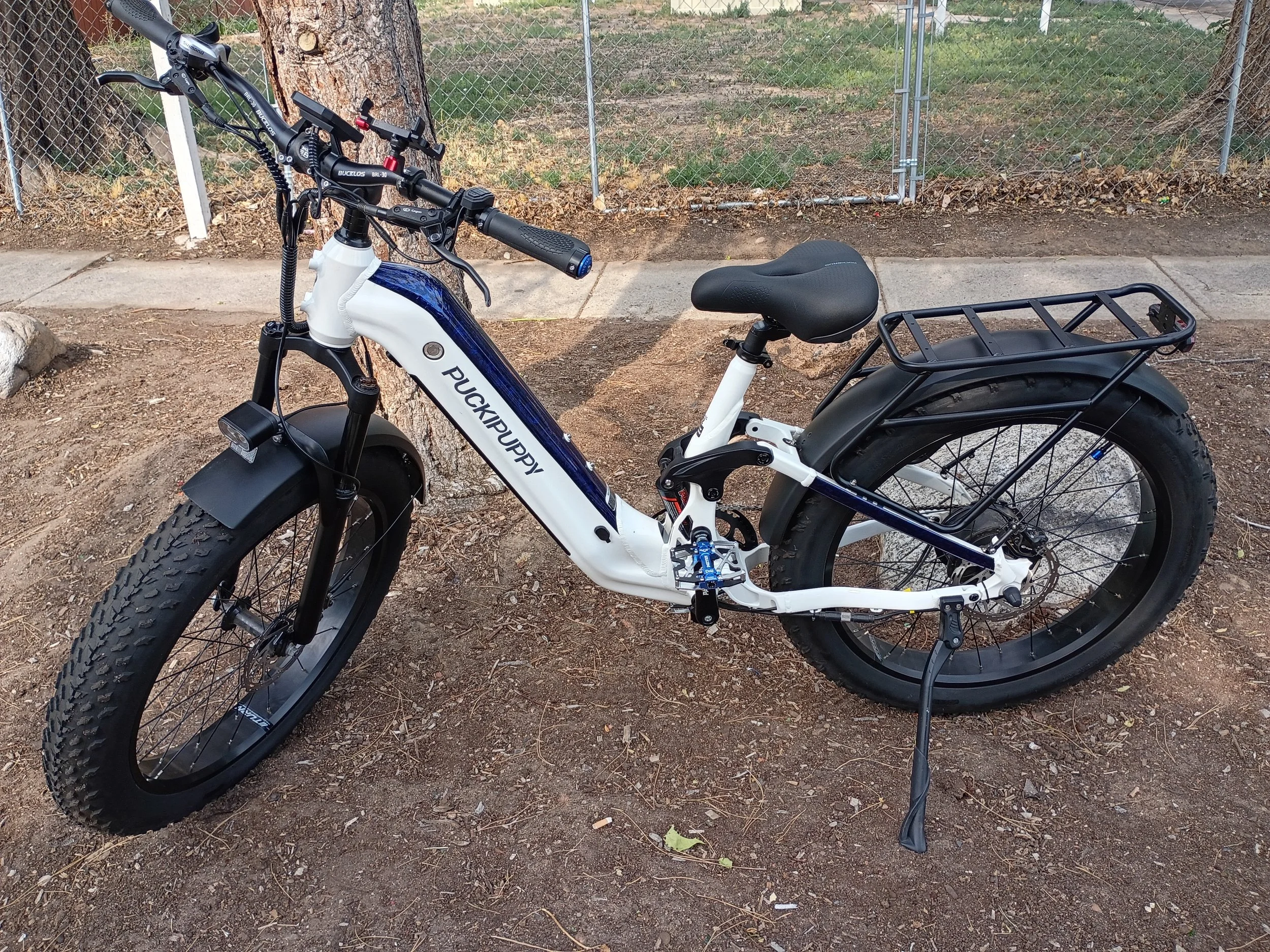 White and black electric bicycle with a rear cargo rack, parked on dirt ground next to a tree and a chain-link fence.