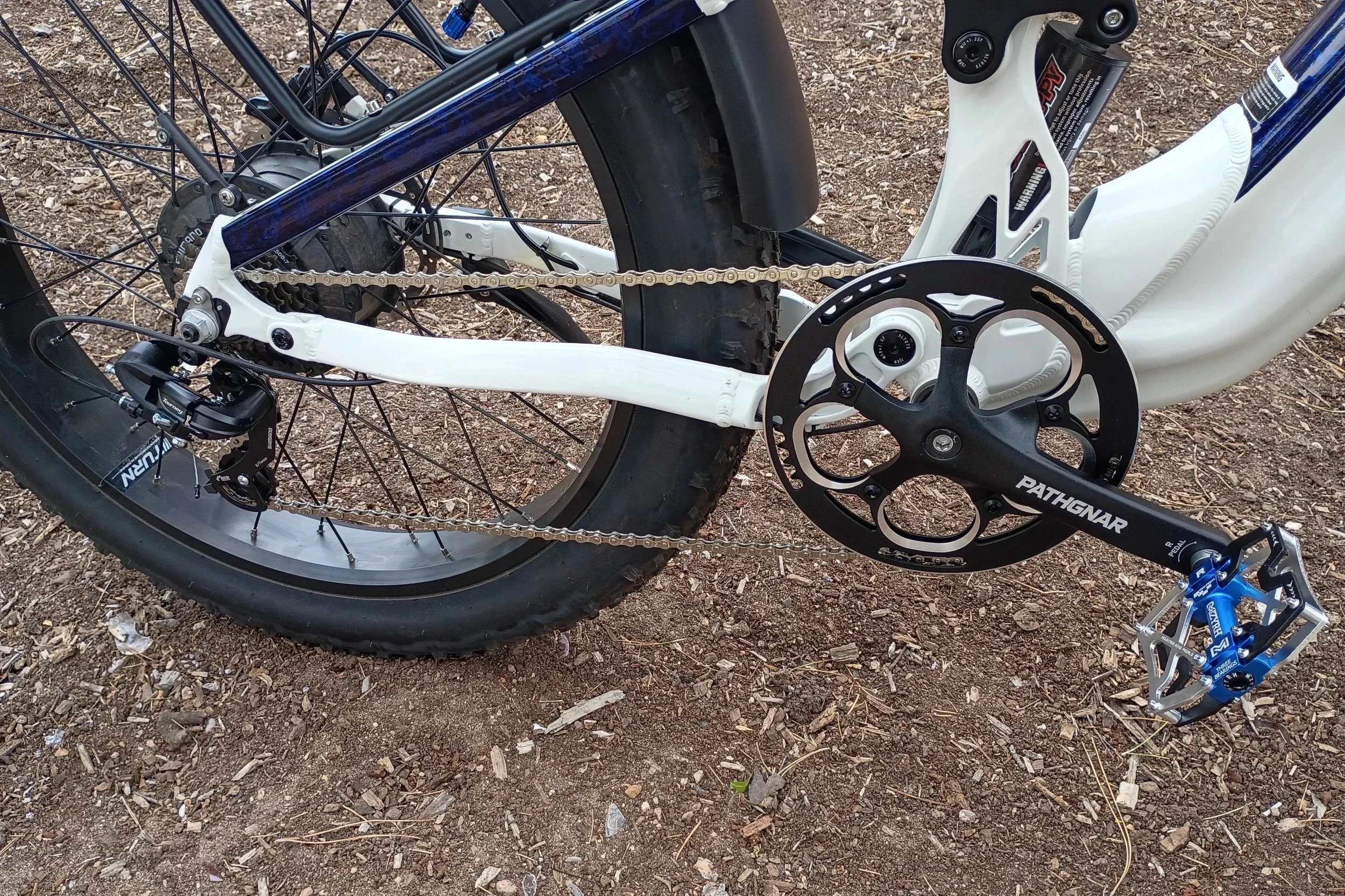 Close-up of a mountain bike's rear wheel, chain, and crankset, with a black tire, white frame, and blue accents, on a dirt surface.