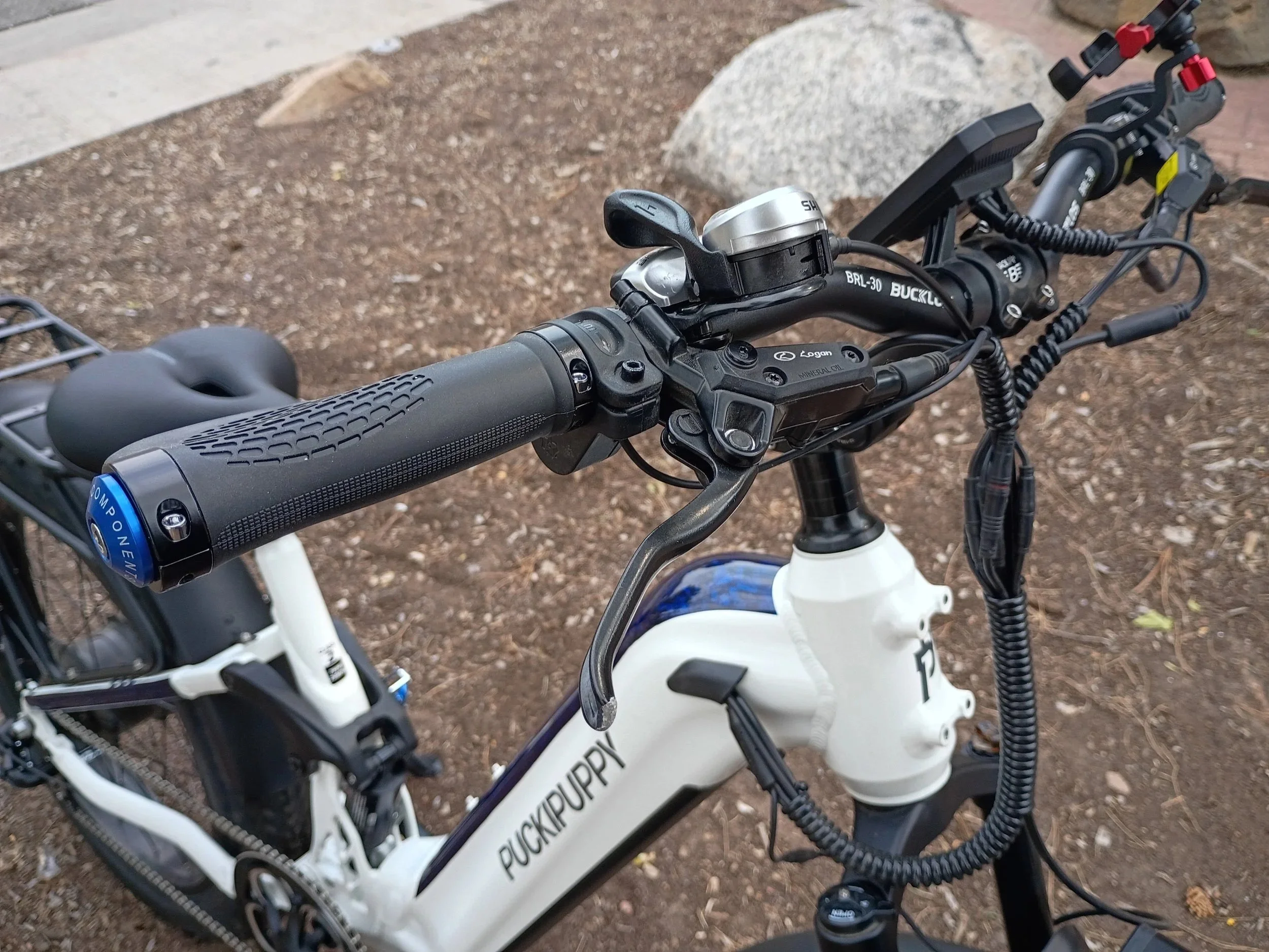 Close-up of a mountain bike handlebar, showing a gear shifter, brake lever, and throttle grip, with a dirt ground and rocks in the background.