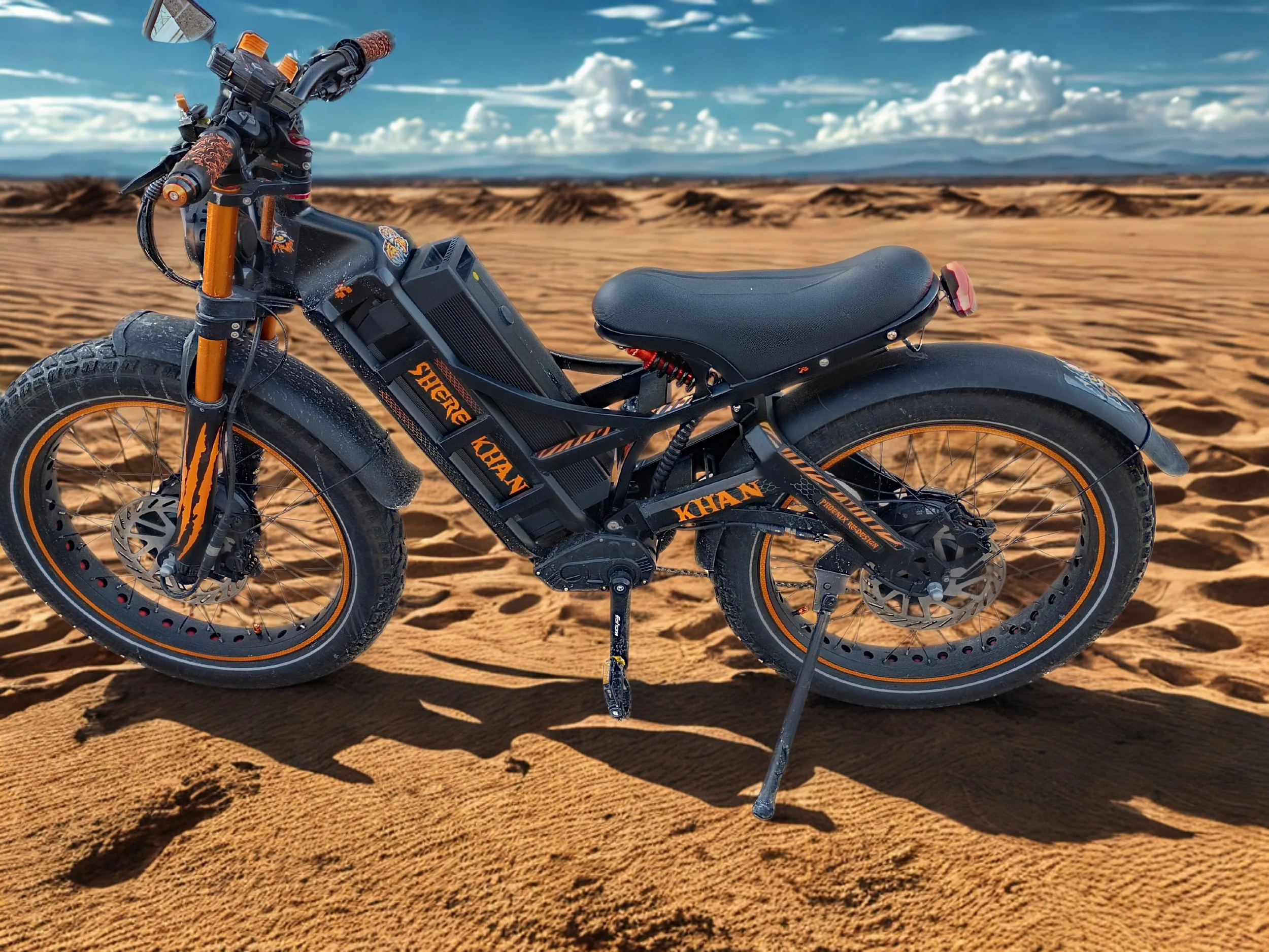 An electric mountain bike with orange accents and stickers is parked on sandy desert terrain under a partly cloudy sky.