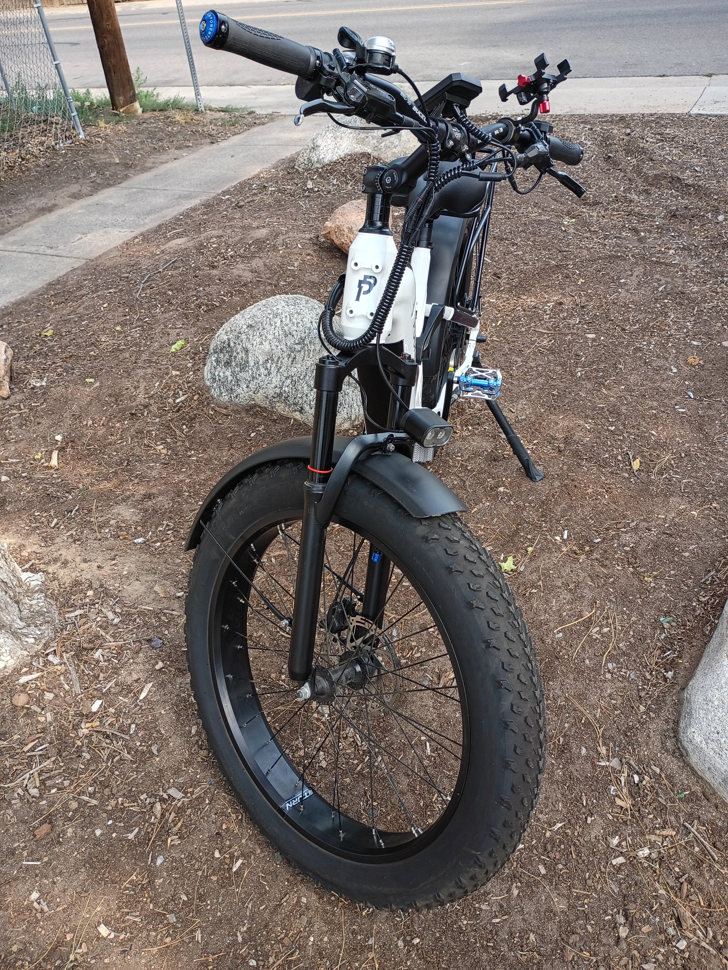 An electric bicycle parked on dirt ground next to a curb and sidewalk, with a city street visible in the background.