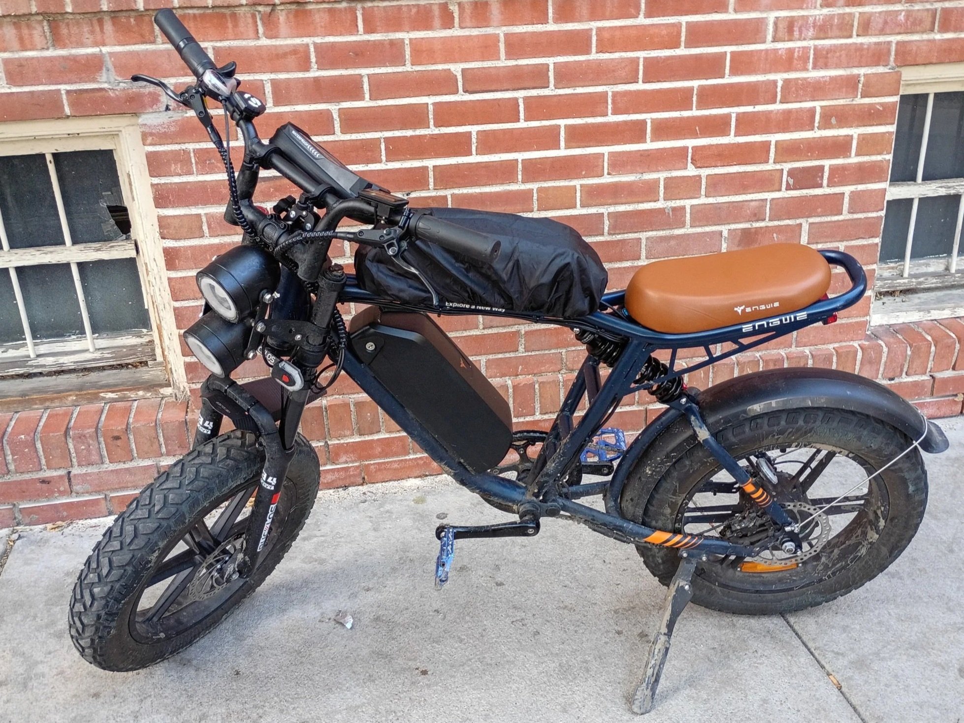 A black electric bicycle with a brown leather saddle and a black handlebar bag, parked in front of a brick wall with two small windows.