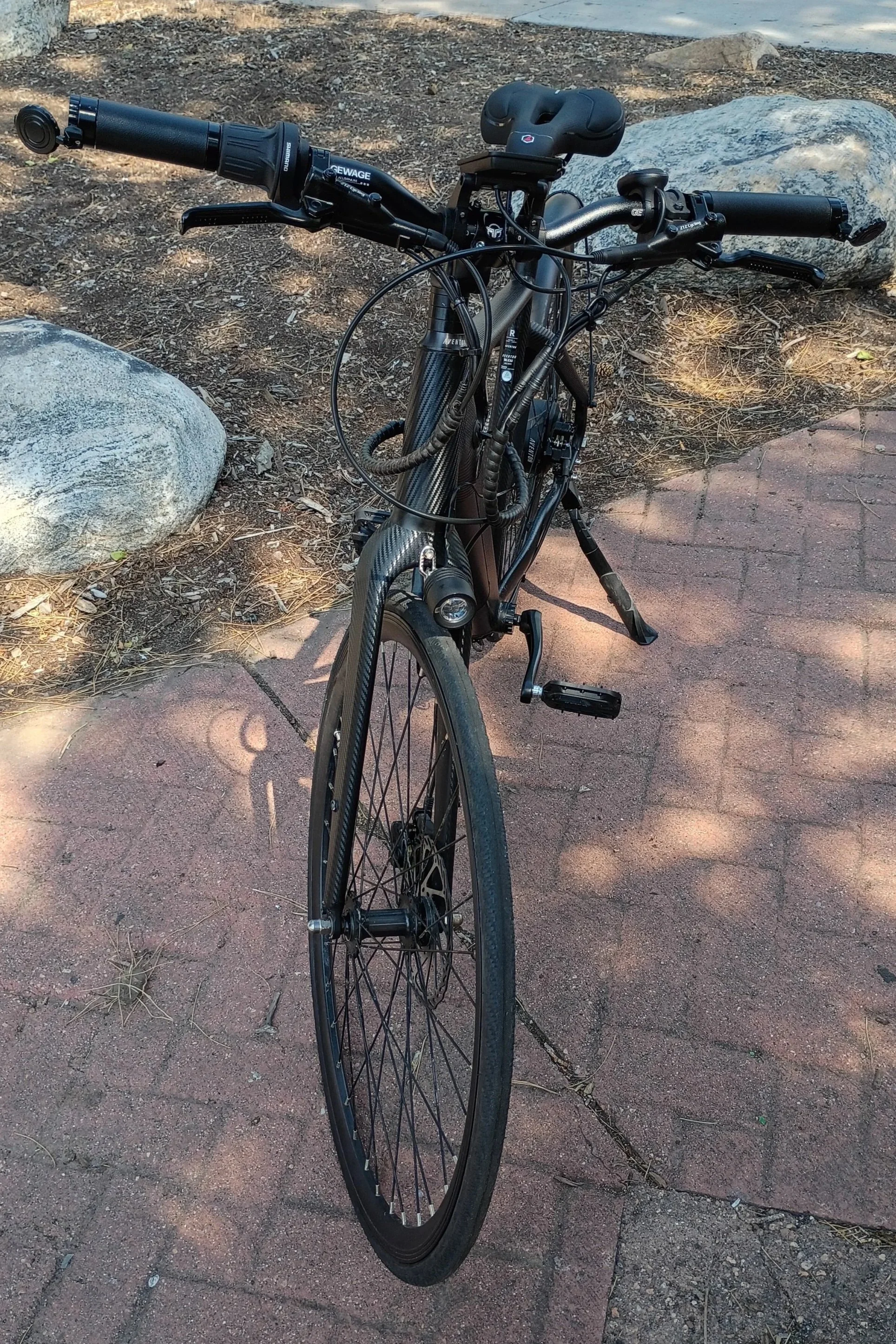 A black mountain bike with a carbon fiber front fork and wide tires, parked on a concrete sidewalk with large rocks and dirt in the background.