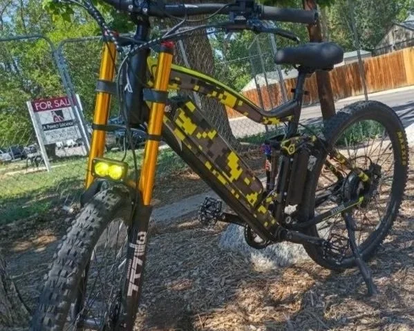 Electric mountain bike with a camouflage pattern, parked on a dirt path near trees and a chain-link fence, with a 'For Sale' sign in the background.