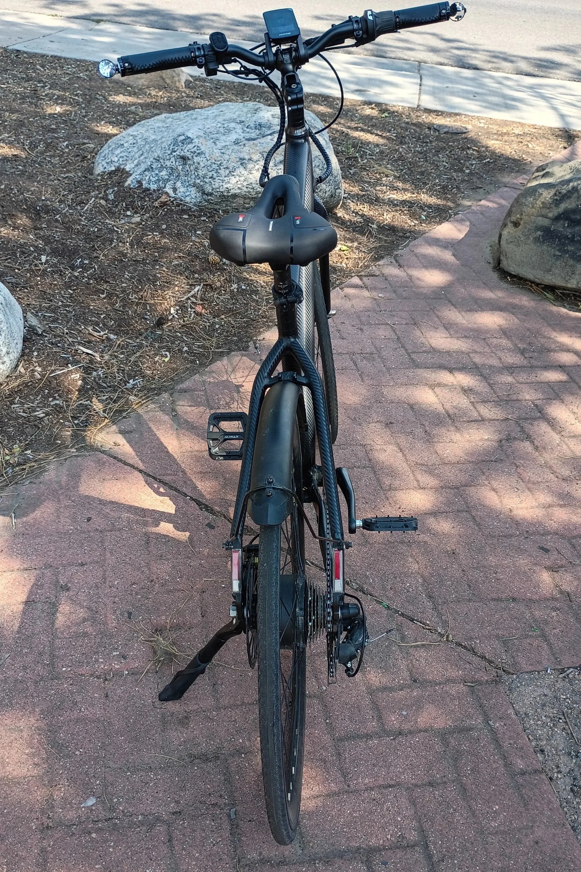 Black mountain bike with a digital device mounted on the handlebars, parked on a brick pathway beside rocky mulch and large rocks.
