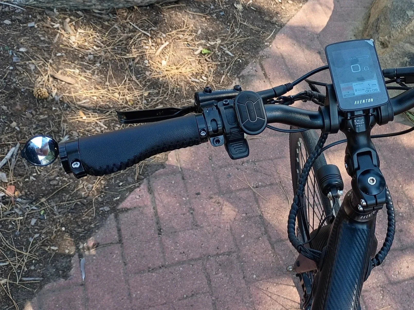 View of an electric bicycle's handlebar, displaying a digital screen, control buttons, a handlebar end bell, and a textured handle grip. The bicycle is positioned on a brick and dirt pathway.