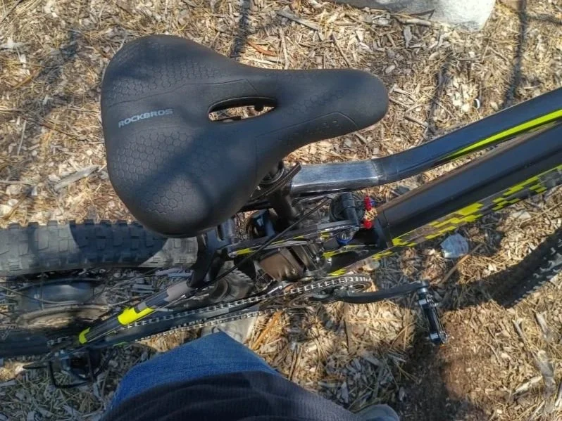 Top-down view of a black and yellow mountain bike with a black RockBros seat, on a dirt trail with leaves and small rocks.