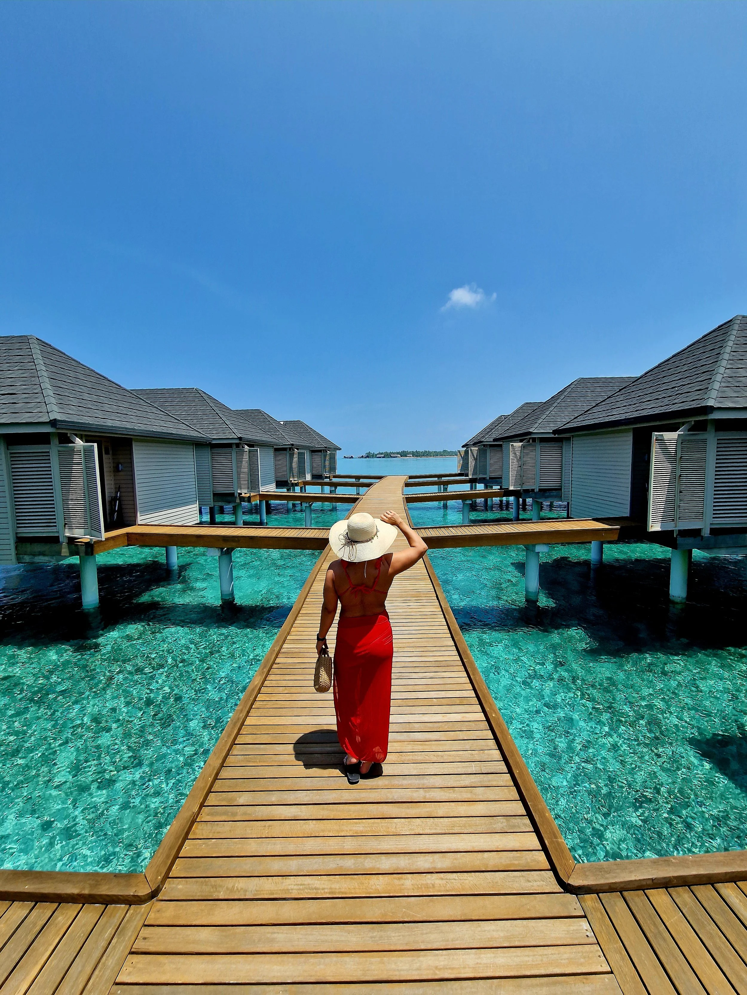 A woman in a red dress and wide-brimmed hat walking on a wooden pier over clear turquoise water, with overwater bungalows and a blue sky in the background.