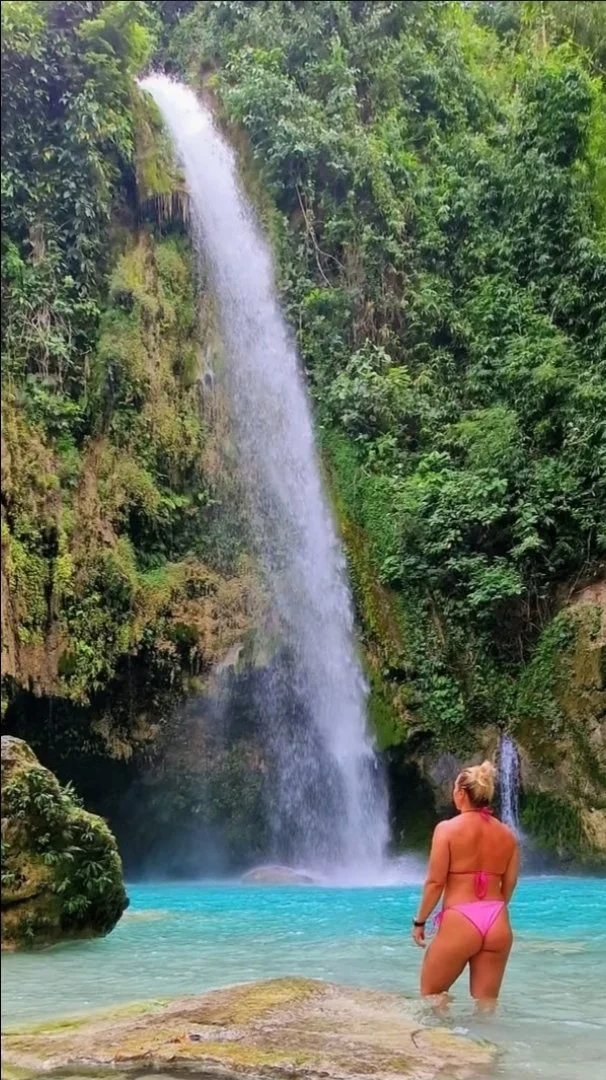 A woman in a pink swimsuit standing in a turquoise pool facing a tall waterfall surrounded by lush green vegetation.