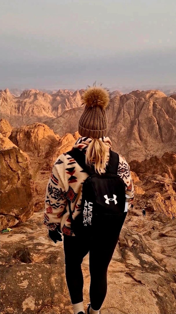 A person in a patterned sweater and knit hat with a pom-pom stands on rocky terrain, facing away, with a mountainous landscape in the background.