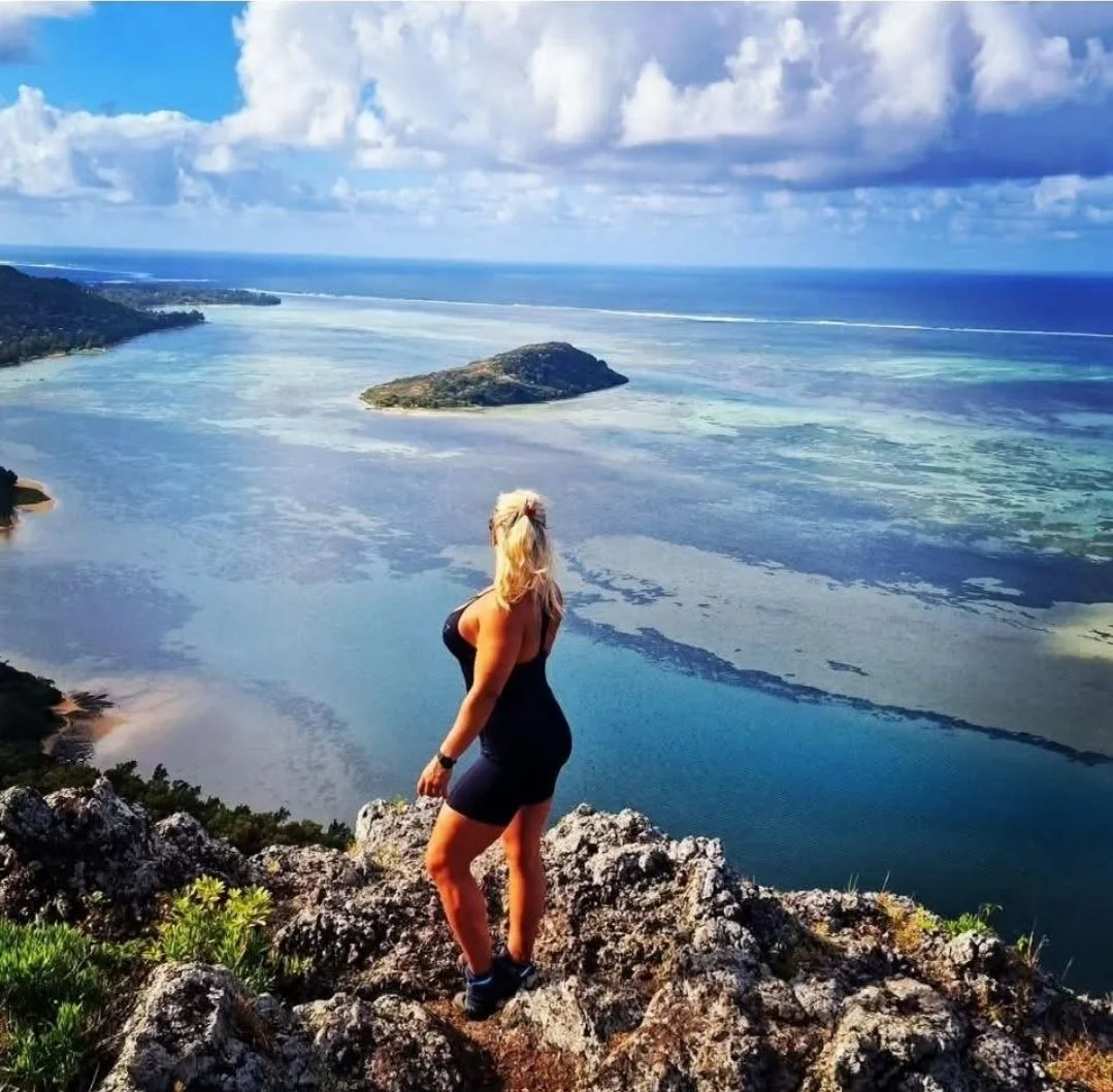 A woman with long blonde hair in a black dress stands on rocky terrain overlooking a large body of water with an island in the distance, under partly cloudy skies.