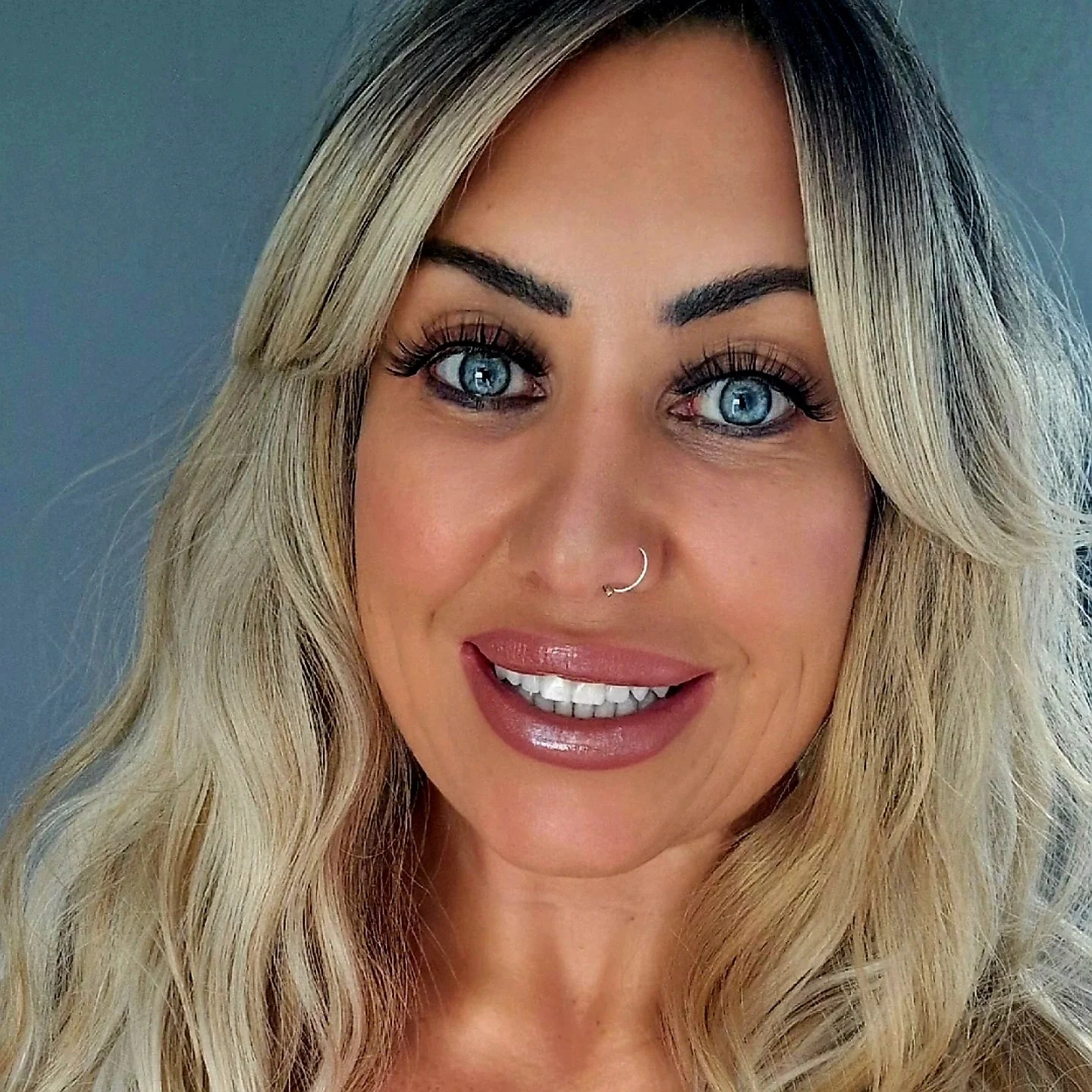 Close-up of a woman with long blonde wavy hair, blue eyes, thick eyebrows, nose piercing, and makeup, smiling with lips slightly parted.