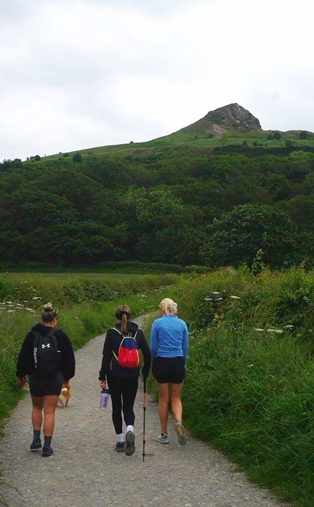 Three women hiking on a trail in a lush green landscape with a rocky hill or mountain in the background.