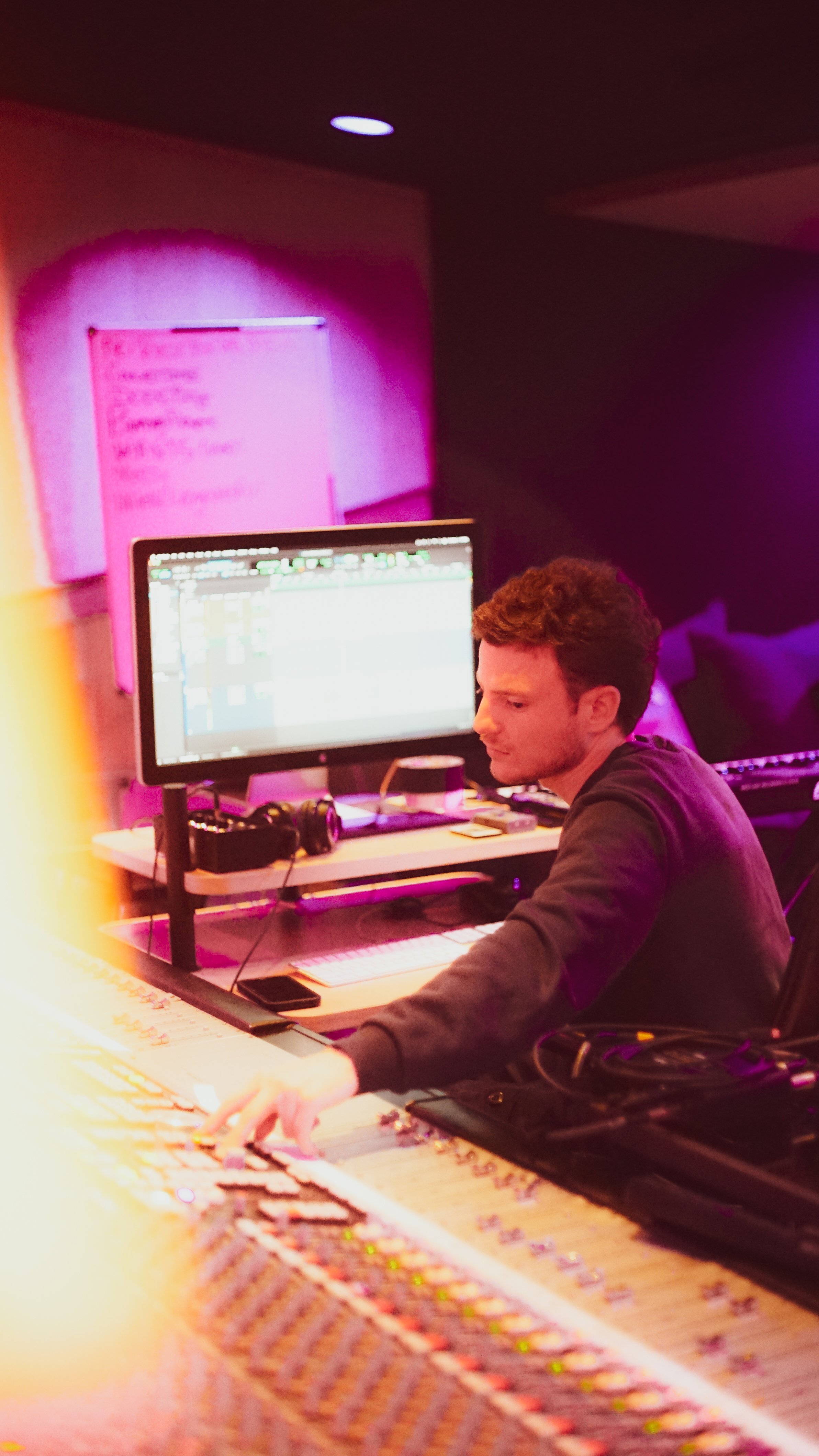 A man working at a music mixing console in a dimly lit room with pink and purple lighting, in front of a computer monitor displaying audio editing software, with a whiteboard on the wall behind him.