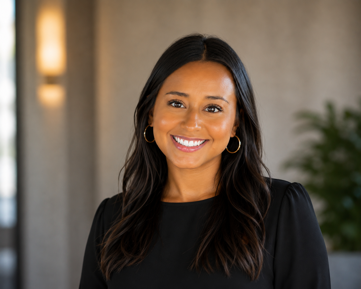 Co-founder and creative director of Constant Light Creative, LLC, Amie Goolsby - with long dark hair, wearing hoop earrings and a black top, smiling indoors with a blurred background.
