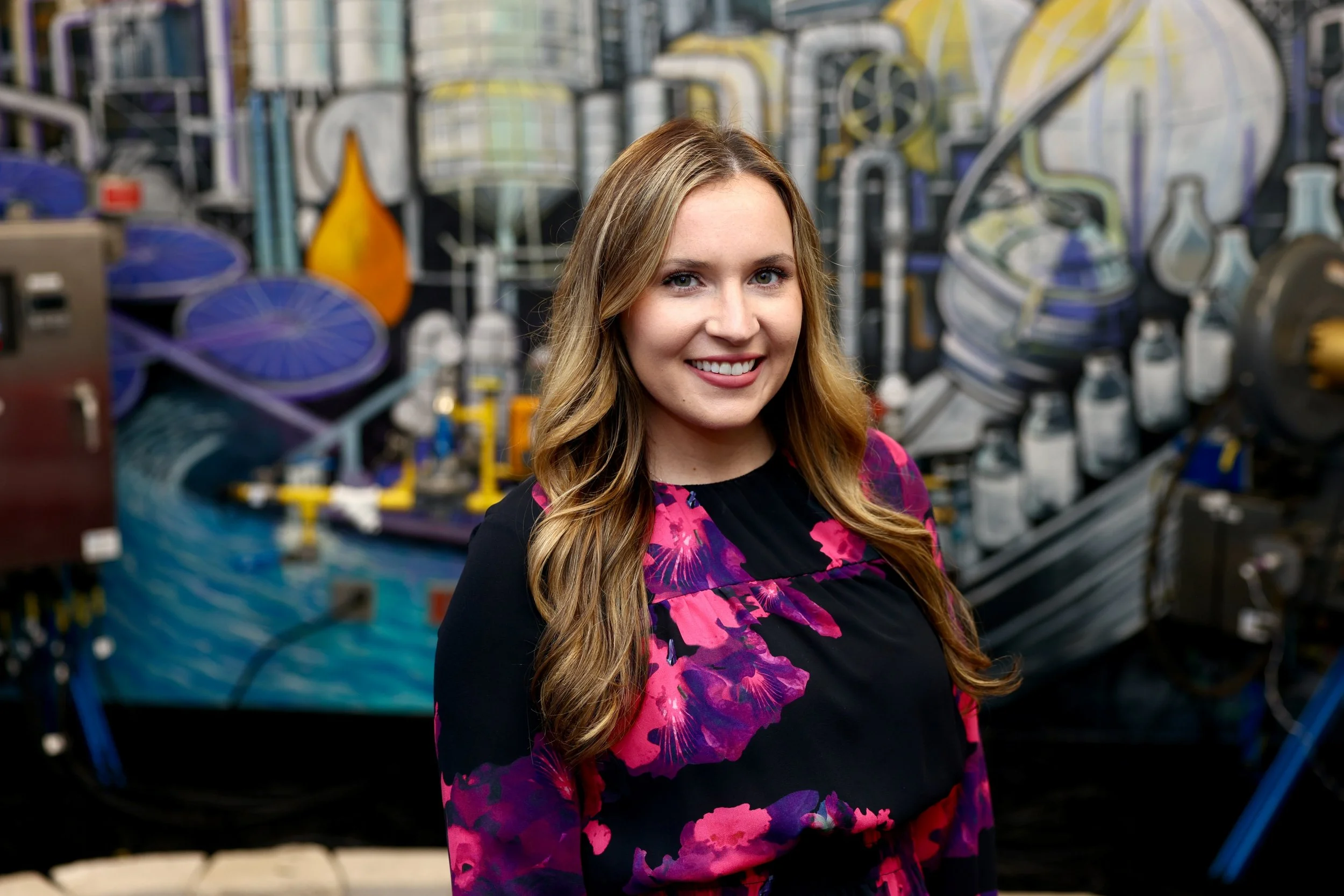 Co-founder and lead strategist at Constant Light Creative, LLC, Shelby Rojas - smiling, with long wavy hair and a colorful floral dress standing in front of a graffiti-covered wall in an urban environment.